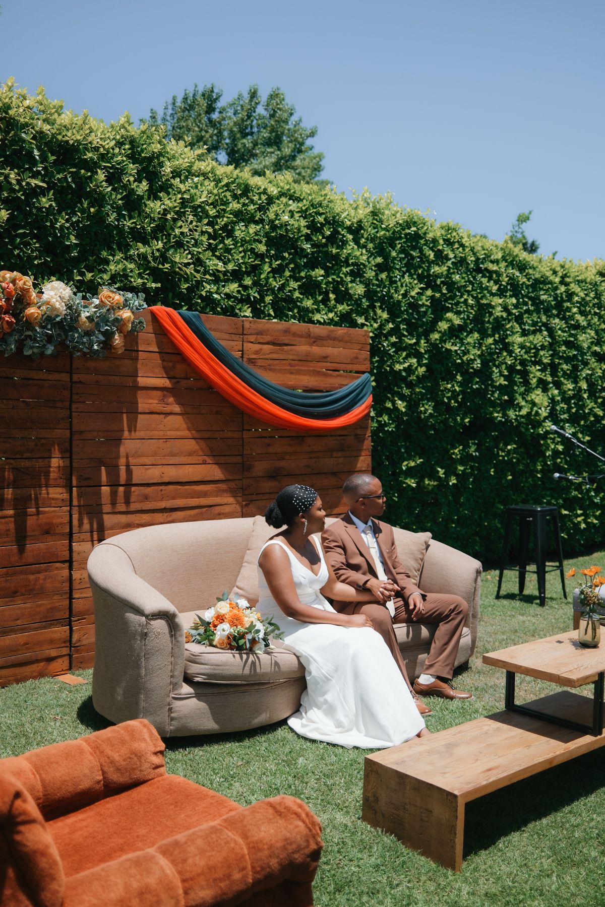 Bride and groom sitting together on a beige sofa in a decorated outdoor garden wedding venue on a sunny day.