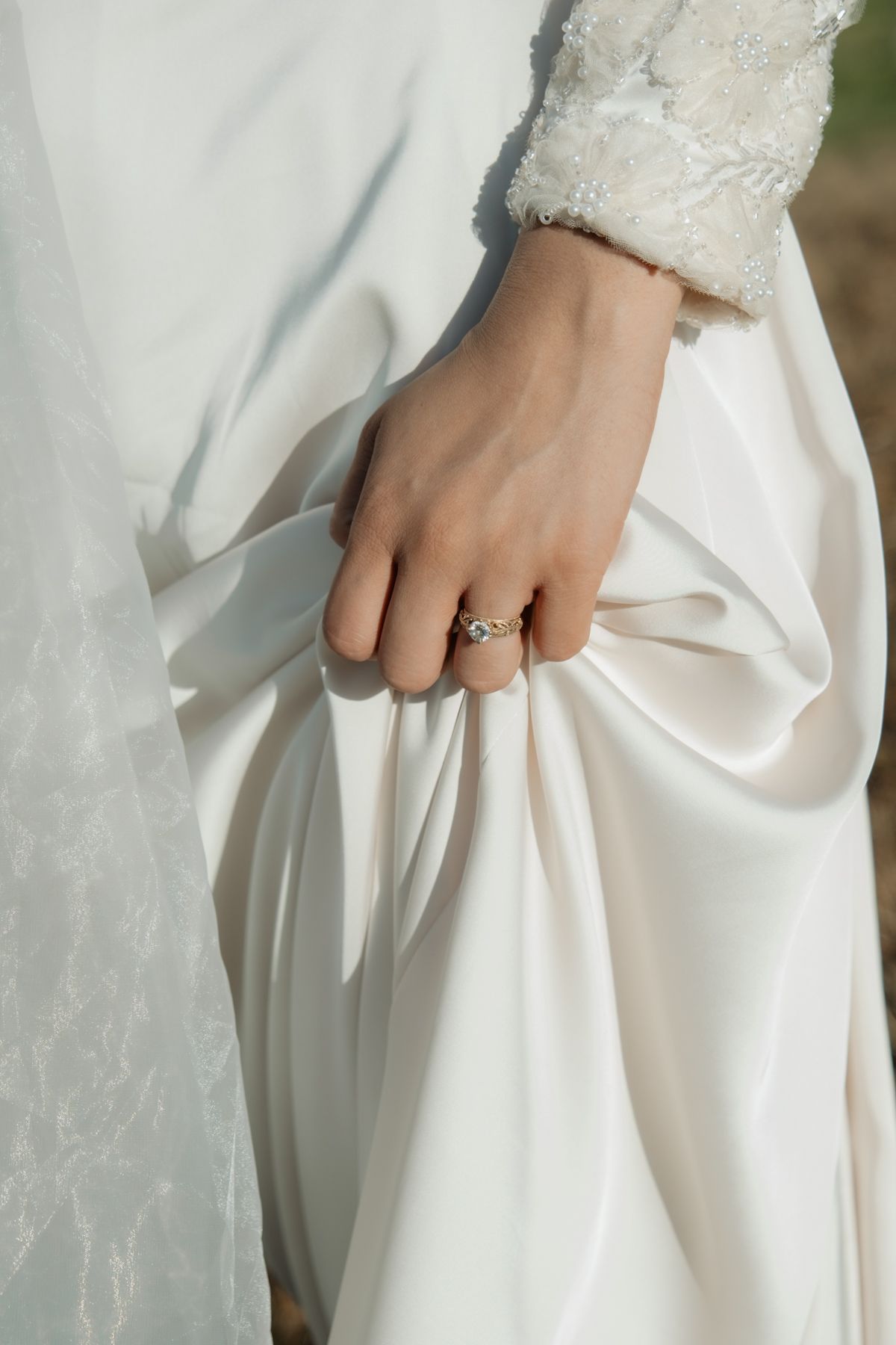 Bride's hand holding white satin wedding dress, wearing a delicate infinity ring, with lace sleeve detail visible.