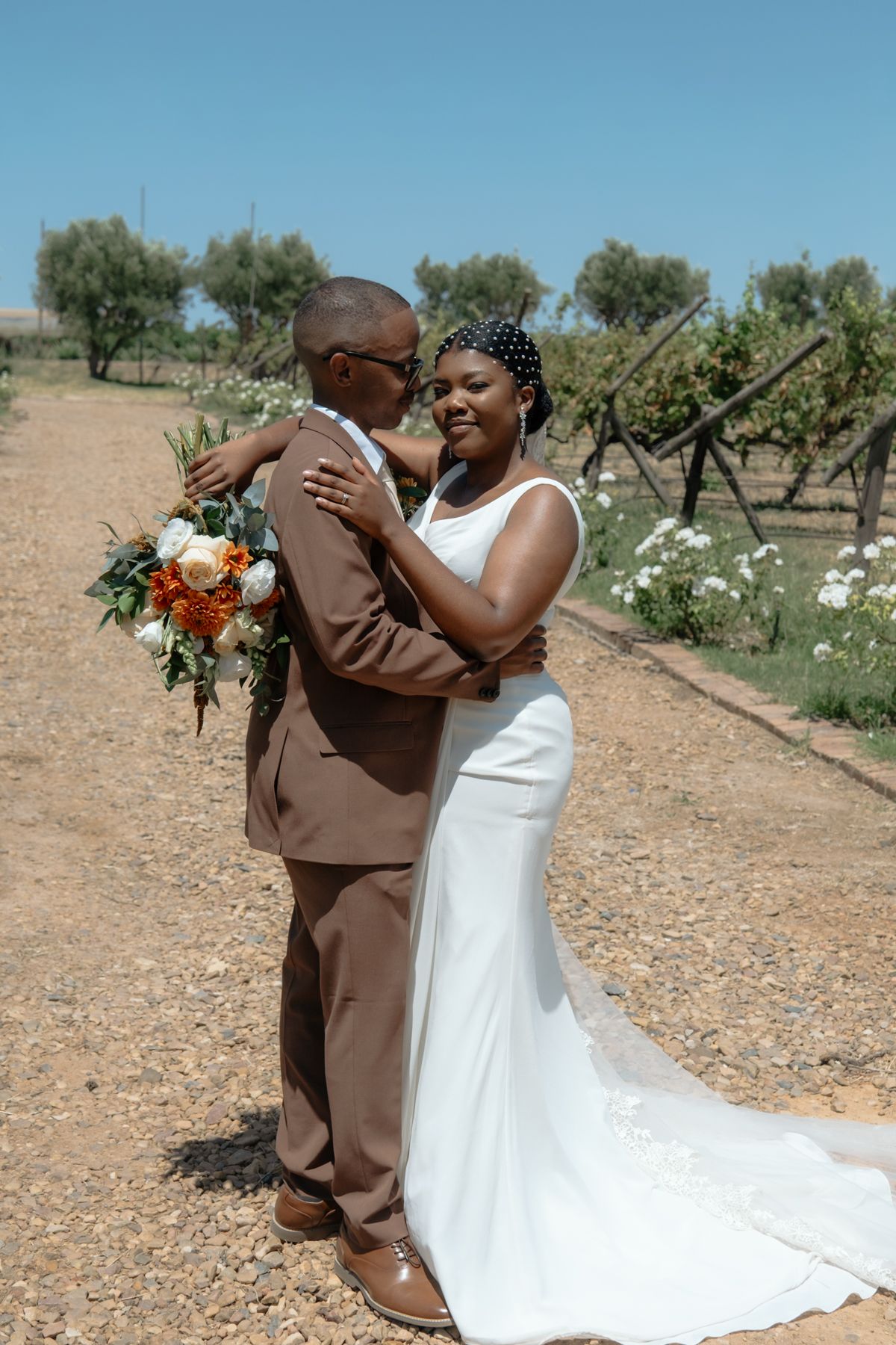 A Black bride and groom embrace outdoors at a vineyard, she in a white gown holding an orange bouquet, he in a brown suit.