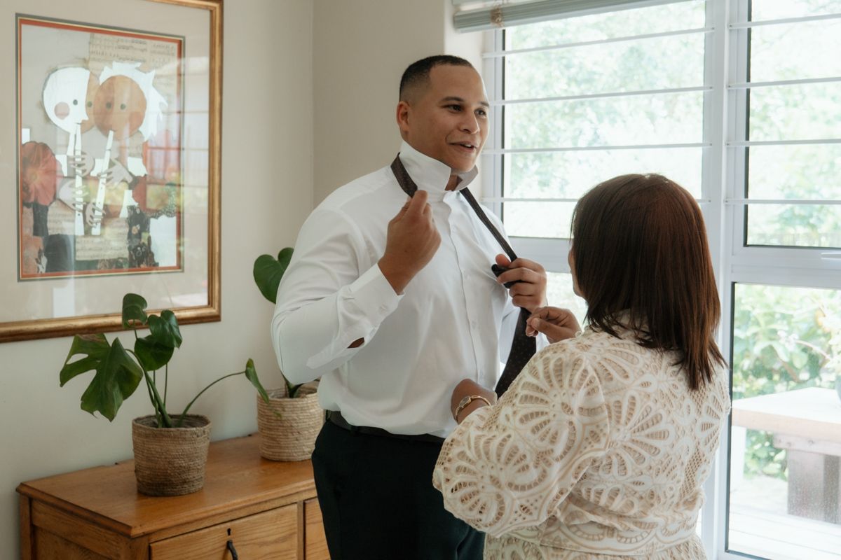 A woman helps a man in a white dress shirt adjust his bow tie while getting ready for a formal event.