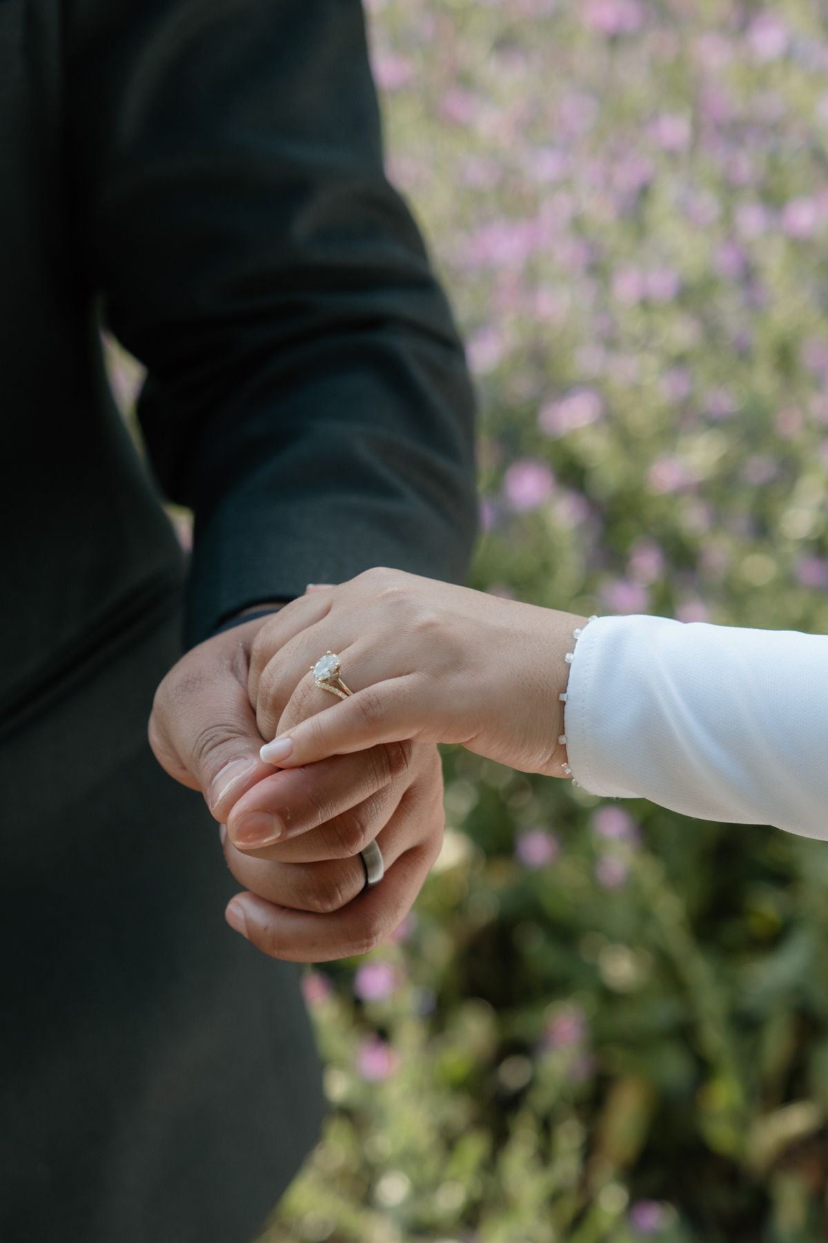 Close-up of bride and groom holding hands showing diamond engagement ring in floral setting.