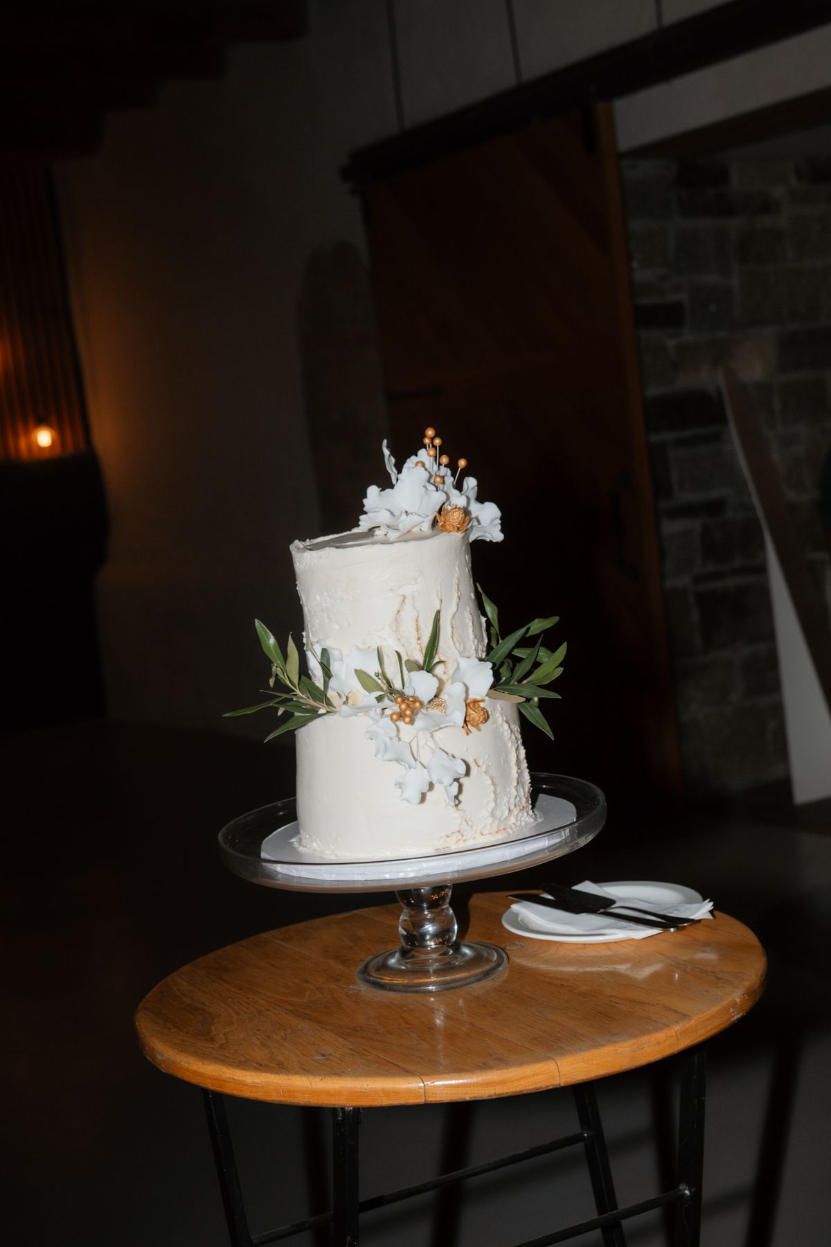 Two-tier white wedding cake decorated with white flowers and greenery displayed on a wooden cake stand.