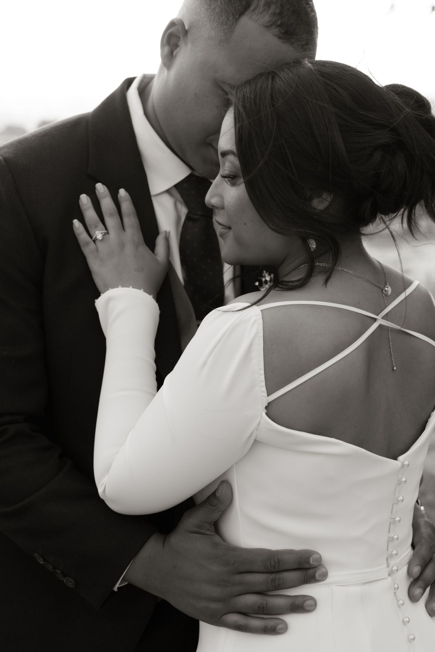 Black and white photo of bride and groom embracing, showing wedding dress back detail and ring.
