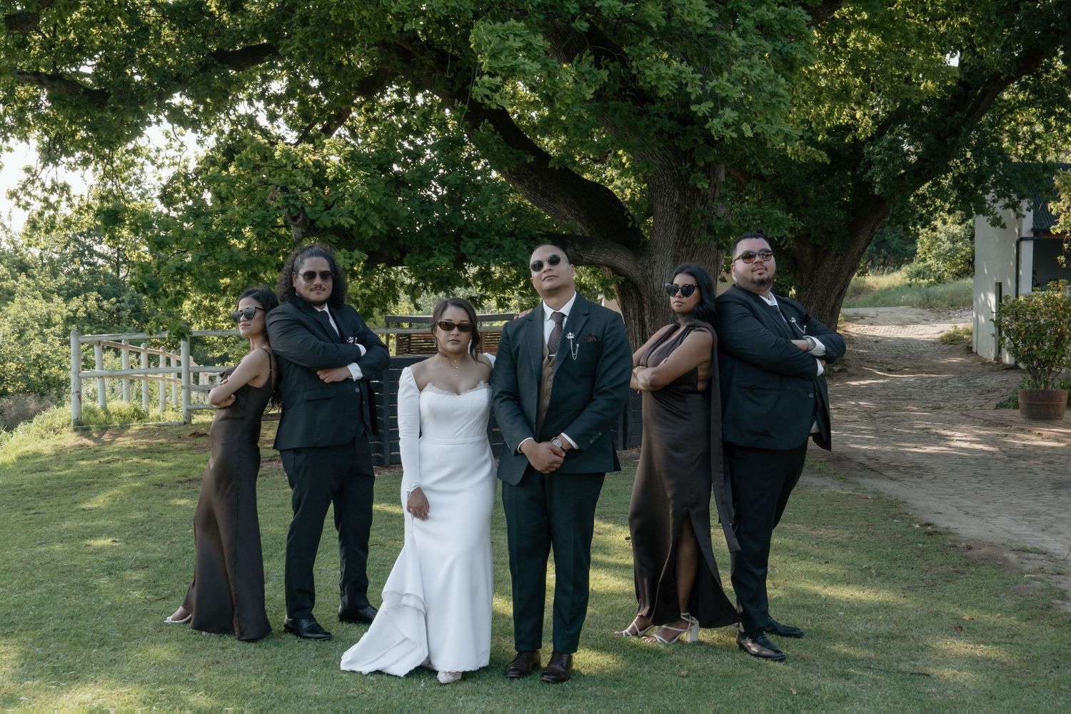 Wedding party posing seriously in sunglasses outdoors under a large tree with bride and groom.