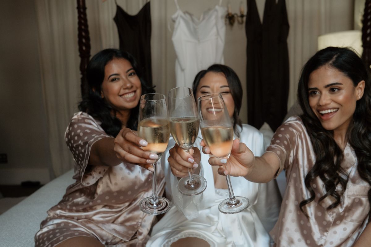 Three bridesmaids in satin robes toasting with champagne glasses while sitting on a bed before the wedding.