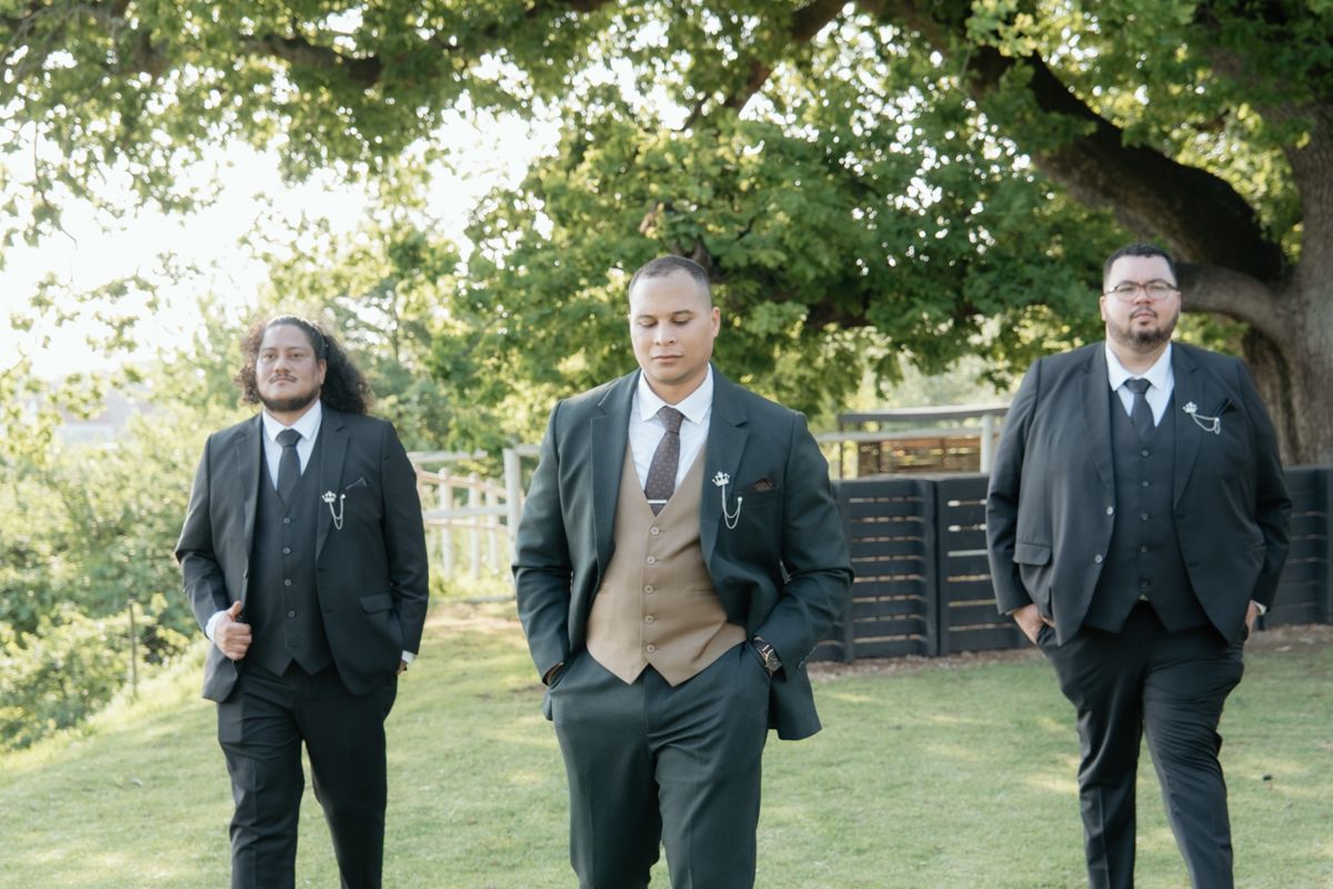 Three groomsmen in dark suits walking confidently across a green lawn under a large tree outdoors.