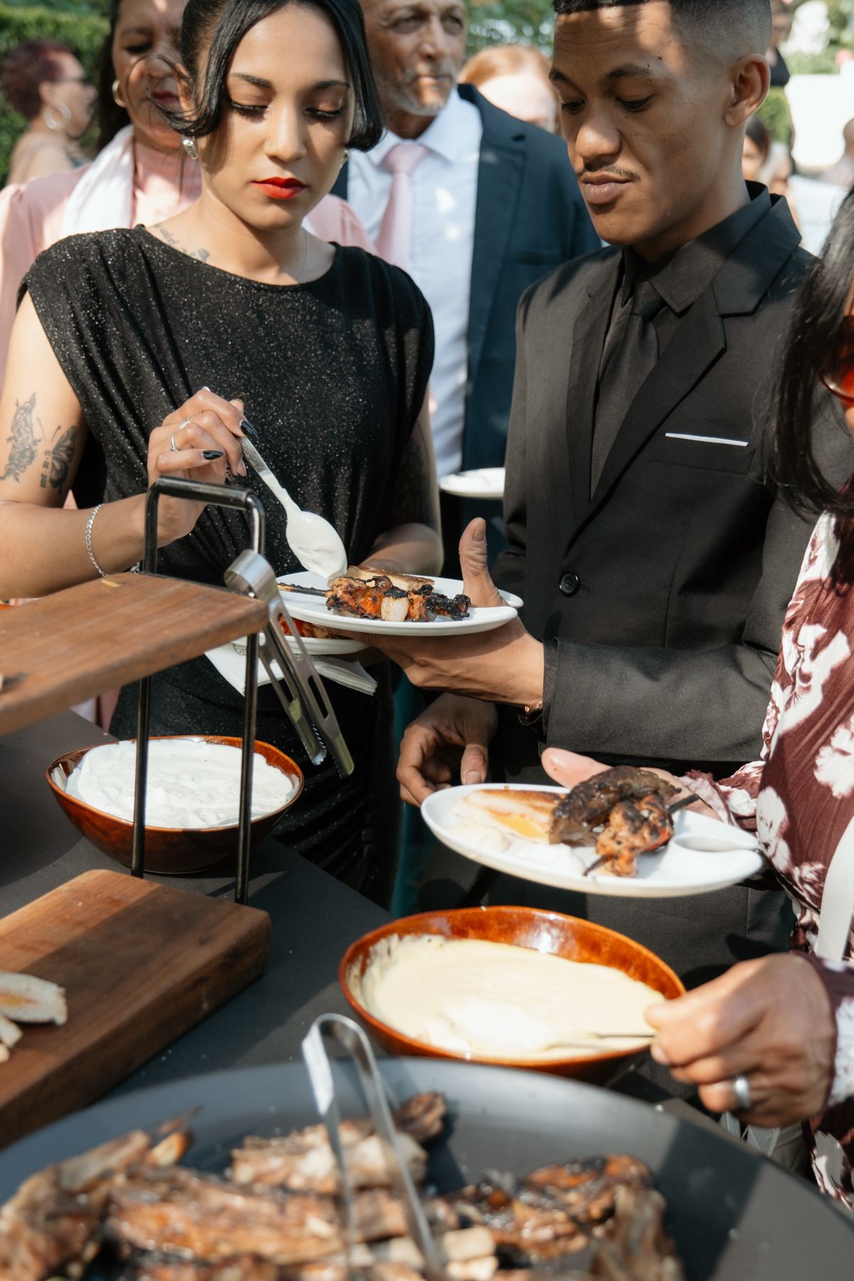 Elegantly dressed wedding guests serving themselves food at an outdoor wedding buffet reception.