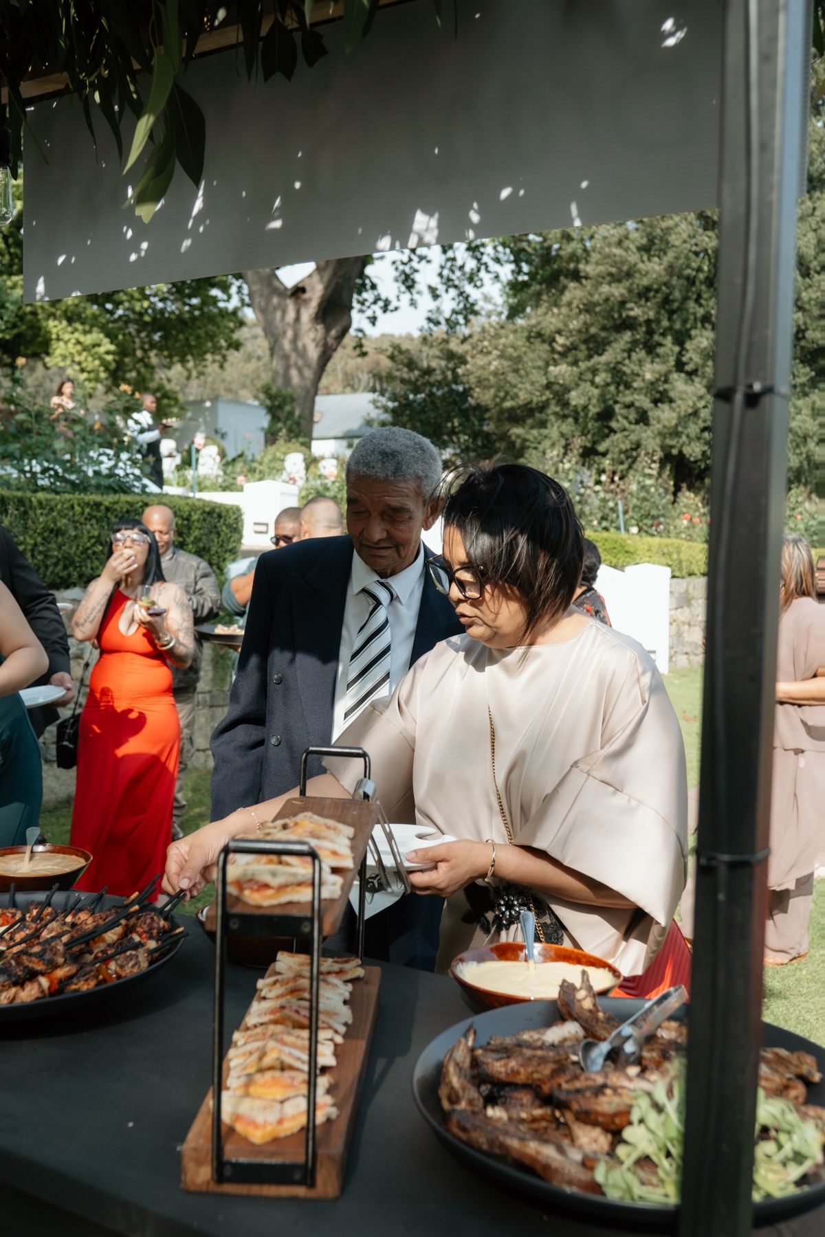 Wedding guests serve themselves at an elegant outdoor buffet spread with grilled meats and dishes.