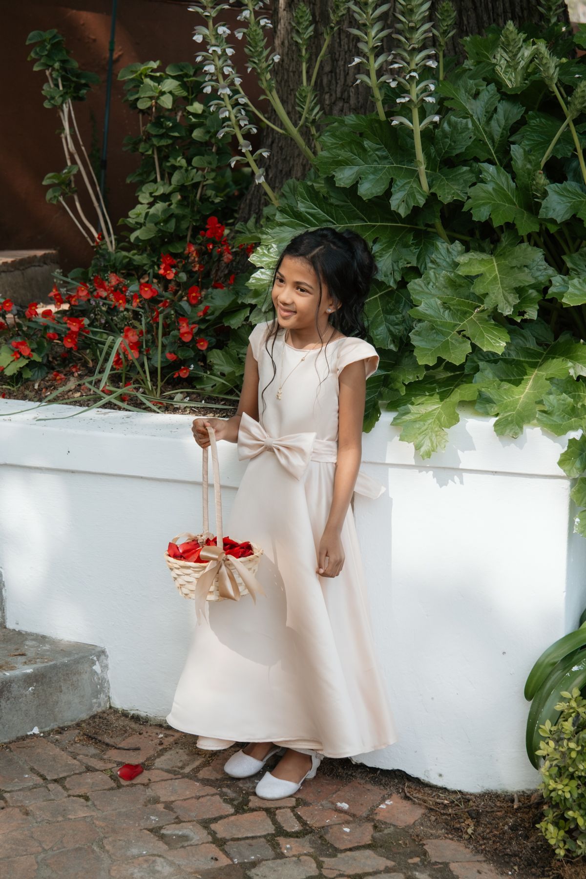 Young flower girl in blush pink dress holding a basket of red rose petals at an outdoor wedding.