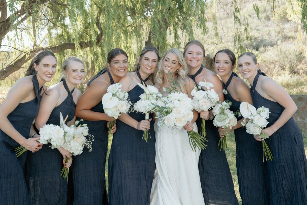 Bride with seven bridesmaids in dark dresses holding white bouquets outdoors.