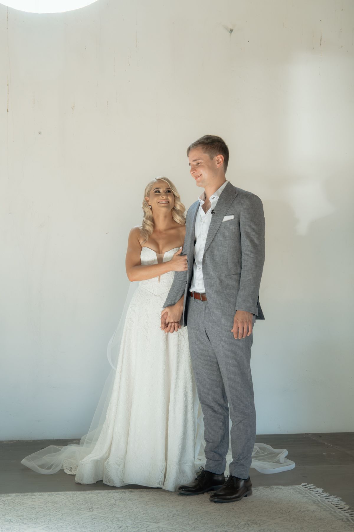 Bride in lace wedding gown and groom in grey suit posing together against a white wall indoors.