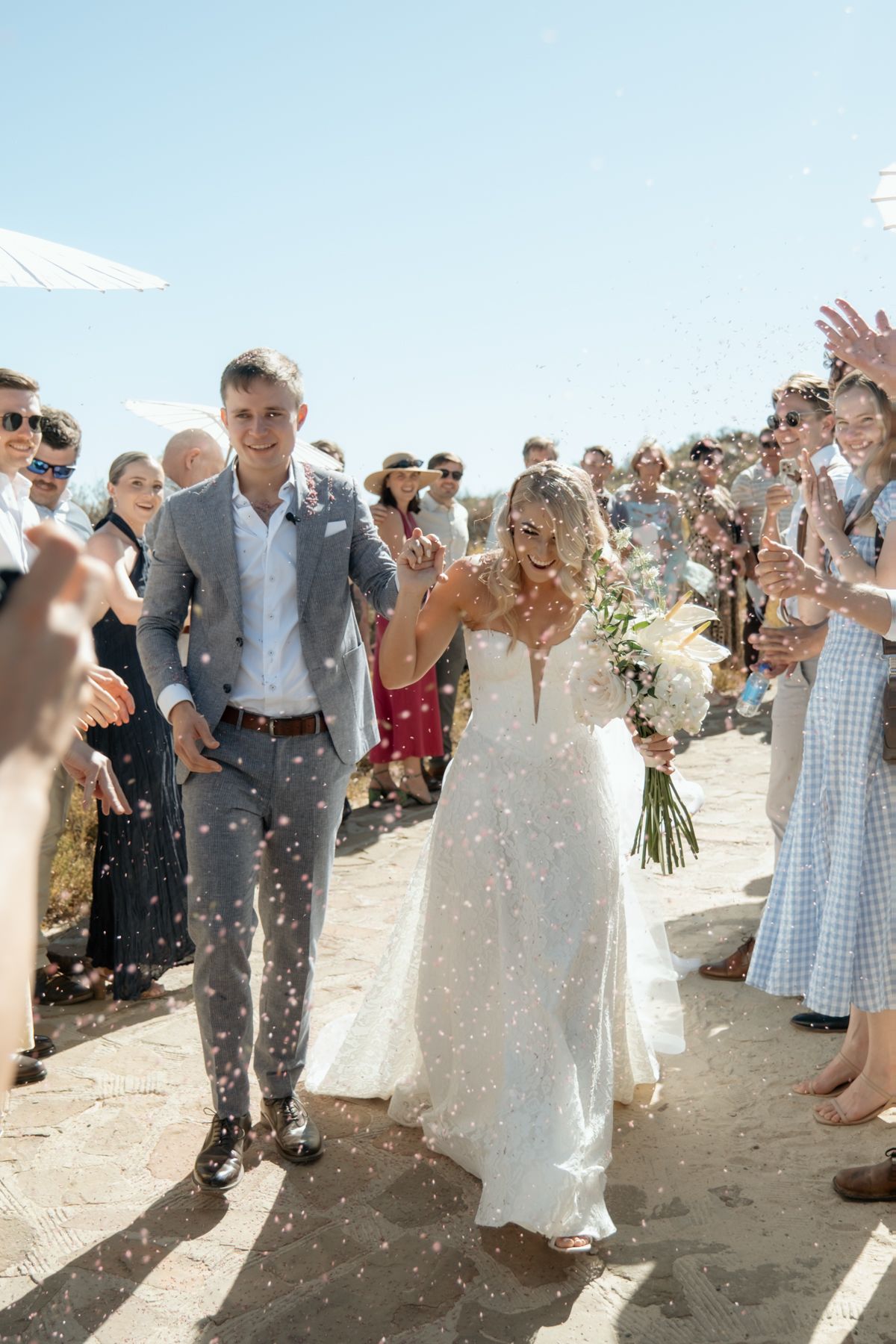 Newlyweds walk through confetti shower as smiling guests celebrate their outdoor beach wedding ceremony exit.
