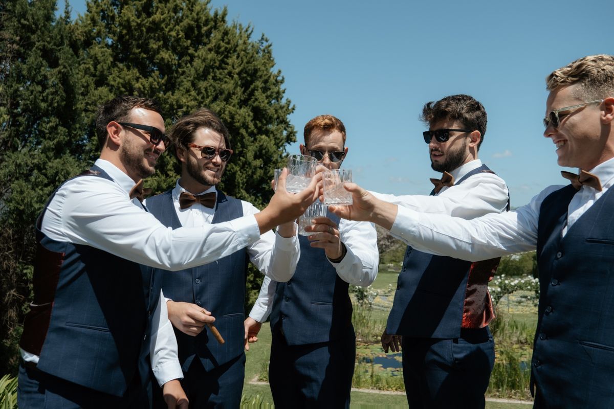 Groomsmen in navy vests and bow ties clinking glasses together in a celebratory outdoor toast.