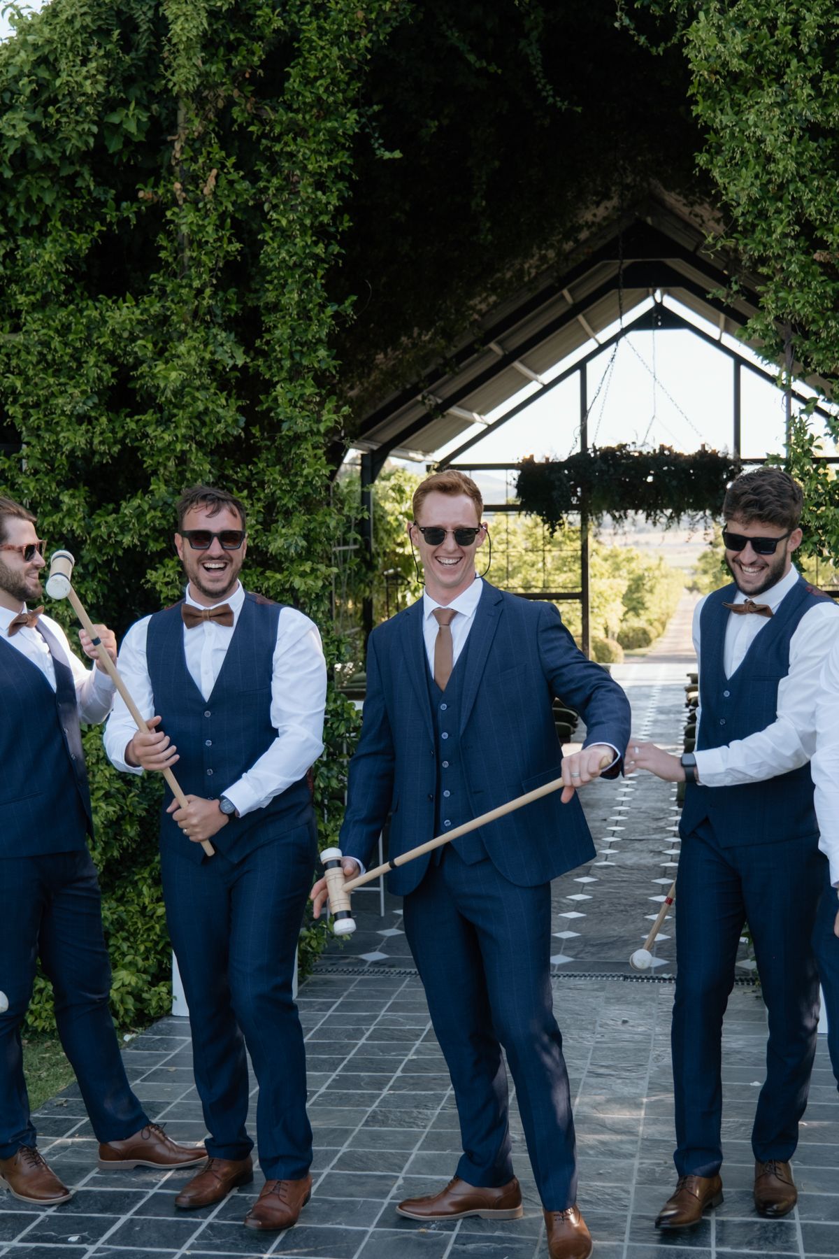 Four groomsmen in navy suits and bow ties pose playfully with canes before an ivy-covered wedding arch.