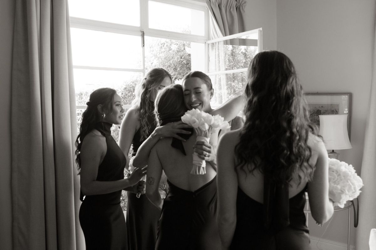 Black and white photo of bride embracing bridesmaids in dark dresses near bright window.