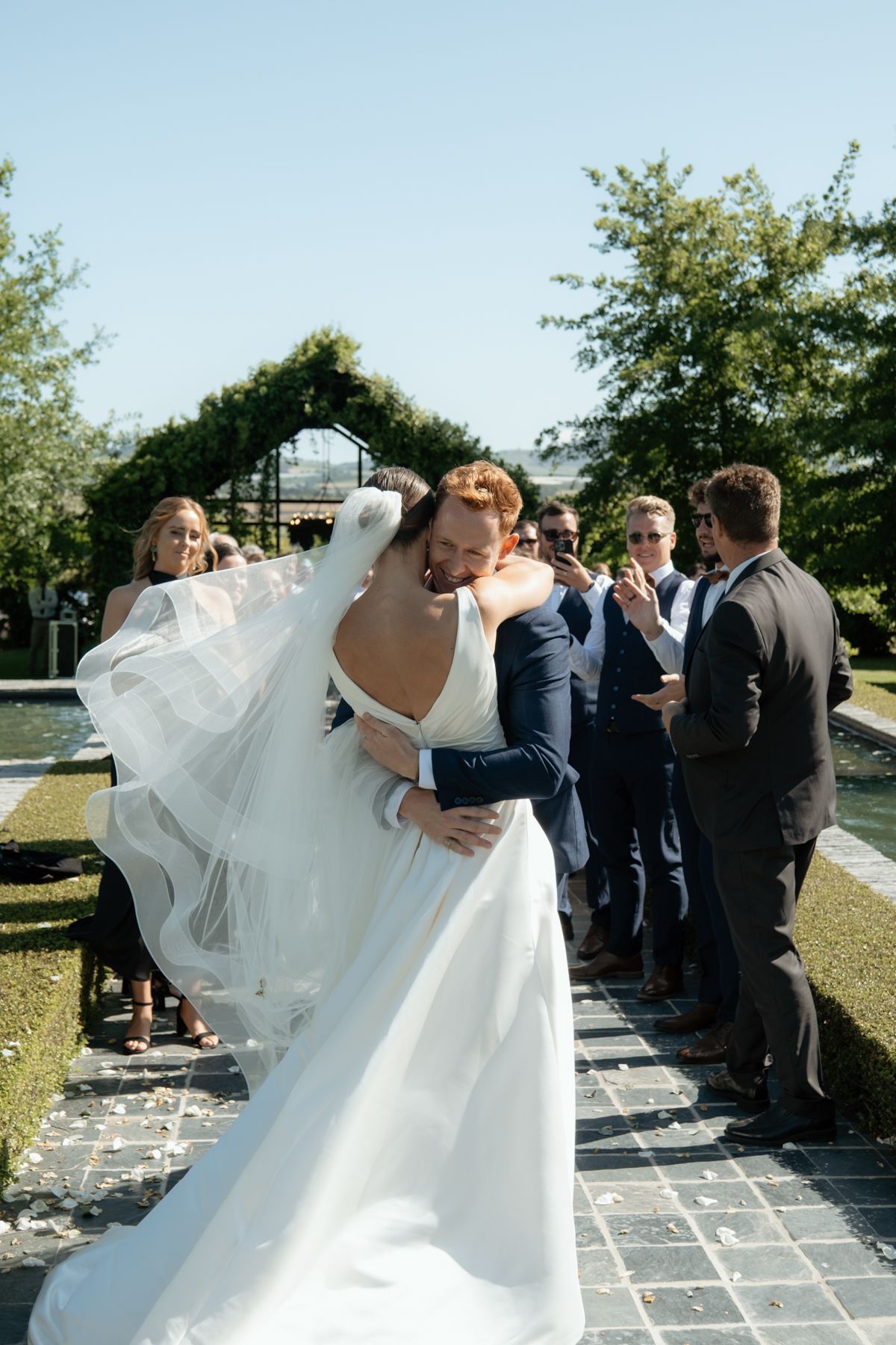 Groom in navy suit lifts bride in white gown for a kiss after outdoor wedding ceremony with guests applauding.