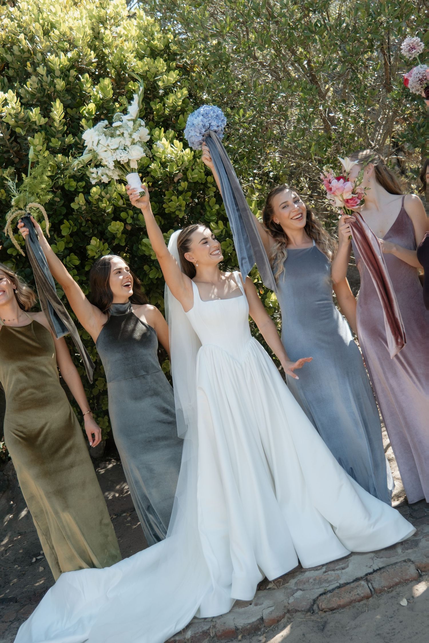 A radiant bride in a white gown celebrates with her bridesmaids in mismatched velvet dresses, raising bouquets joyfully.