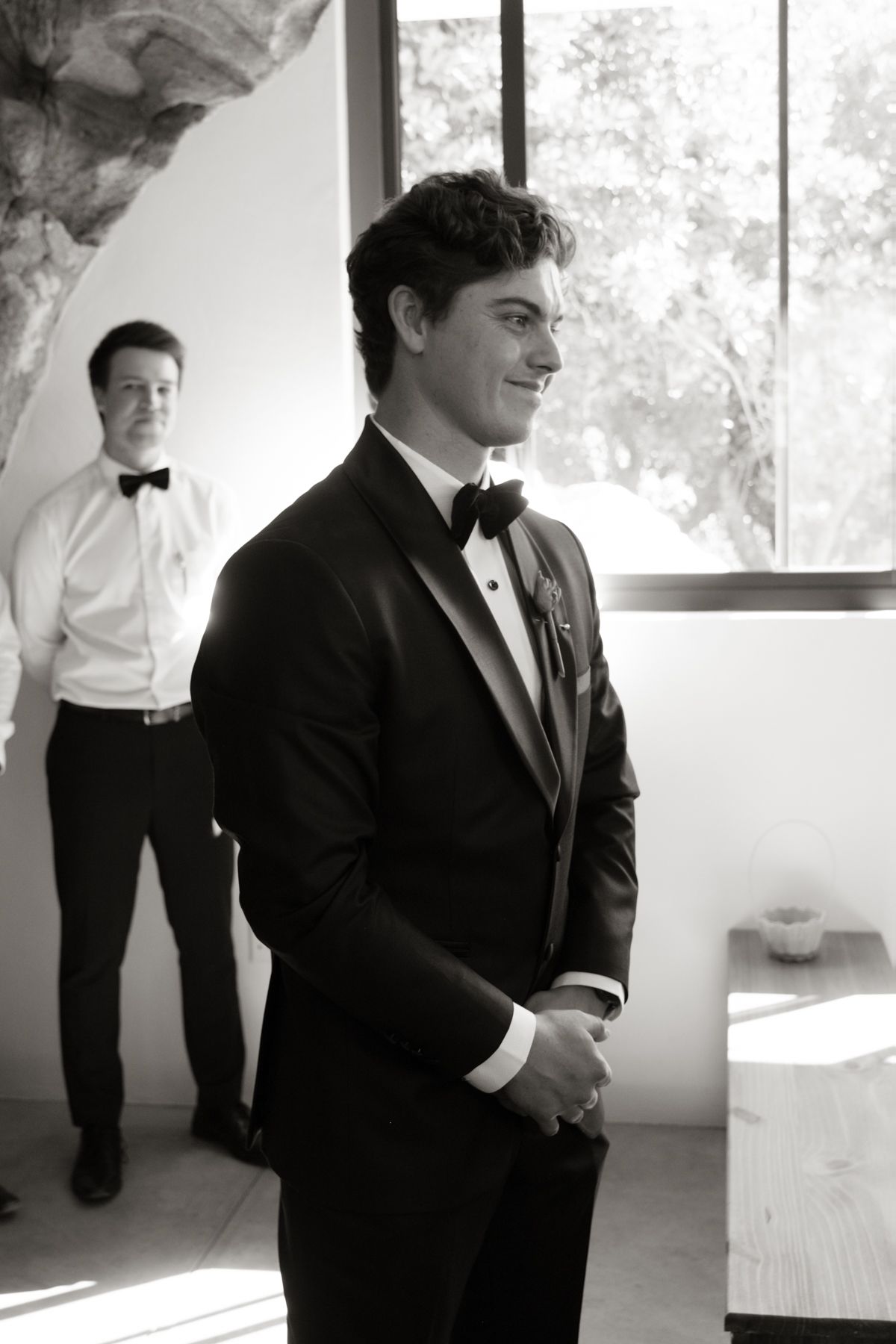 Groom in black tuxedo smiling during wedding preparations, with groomsman in background, black and white photo.