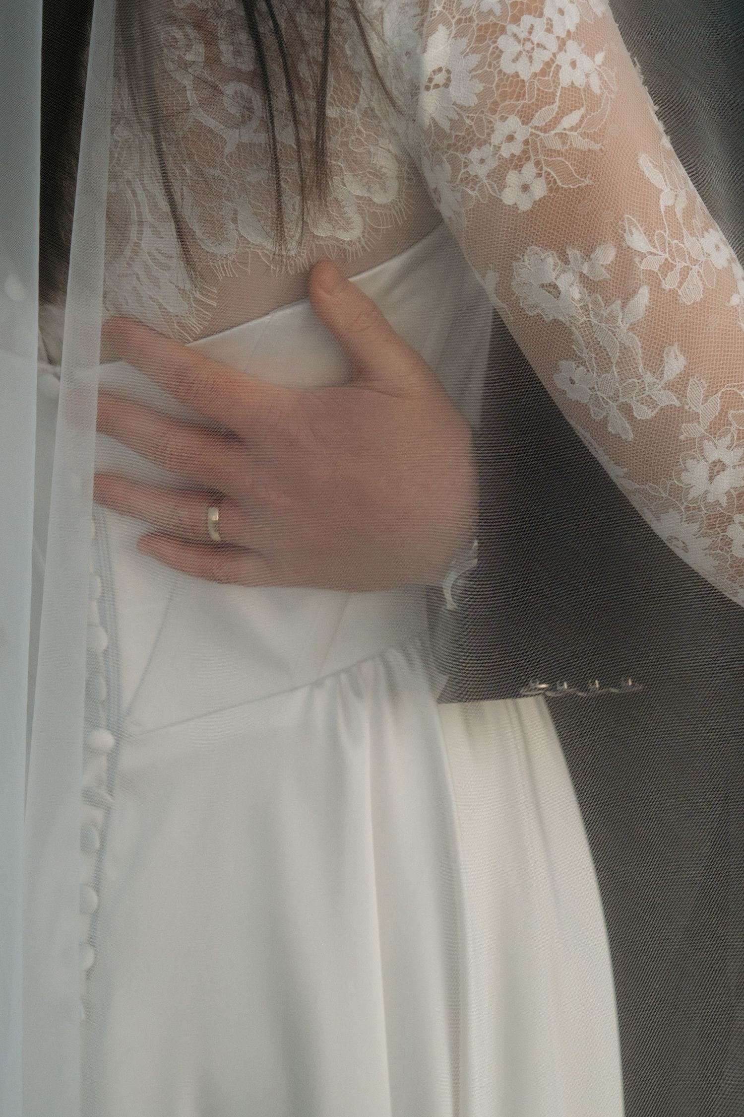 Close-up of groom's hand resting on bride's lace wedding dress waist beneath flowing veil.