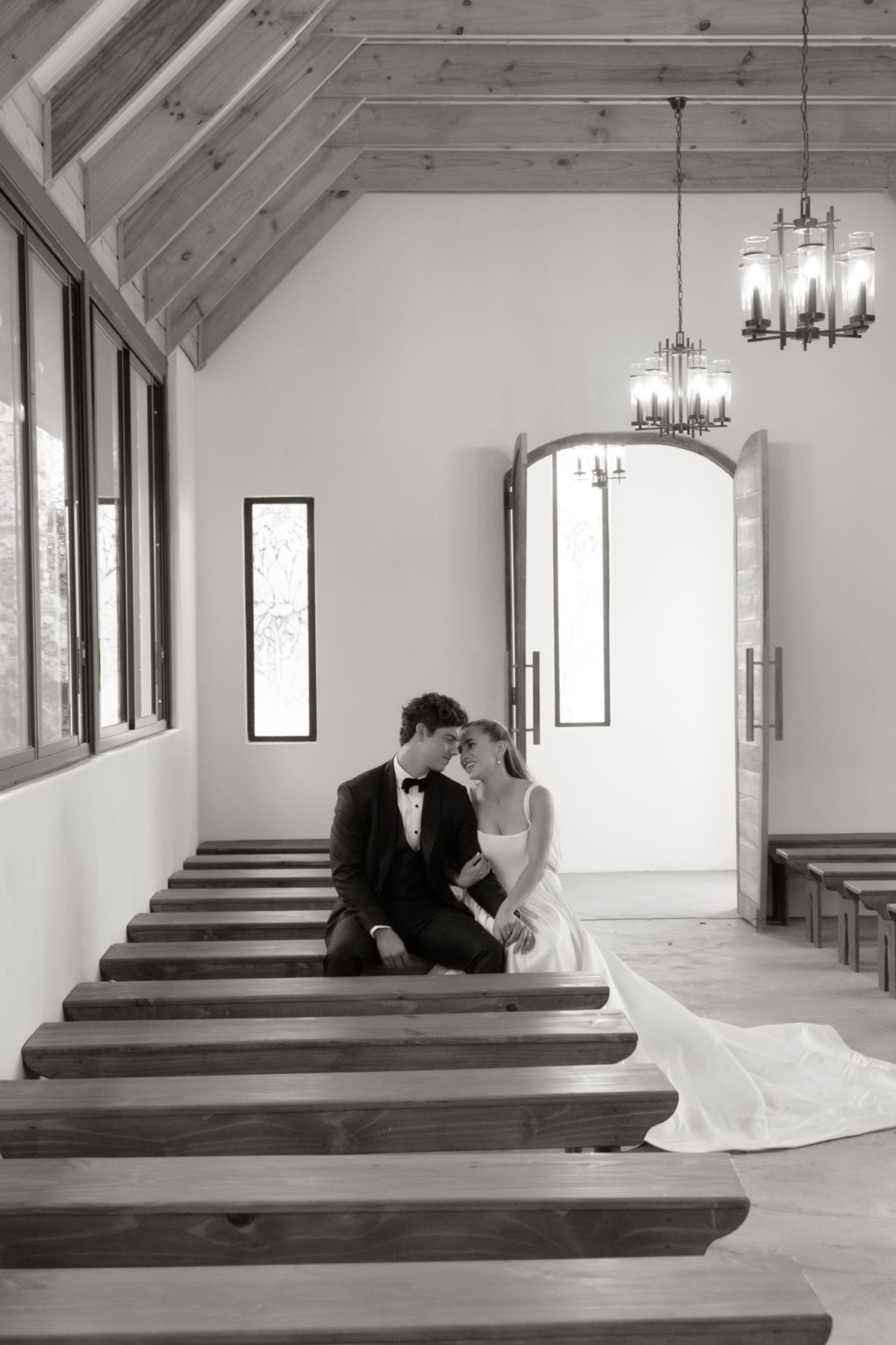 Black and white image of bride and groom sharing intimate moment on chapel pew with chandeliers above.