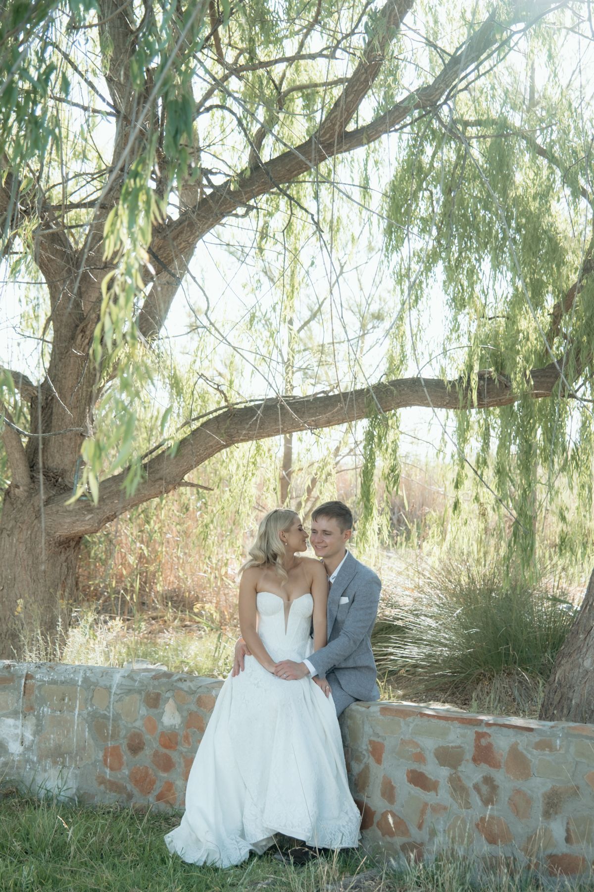 Bride and groom sitting together against stone wall under large willow tree in natural setting.