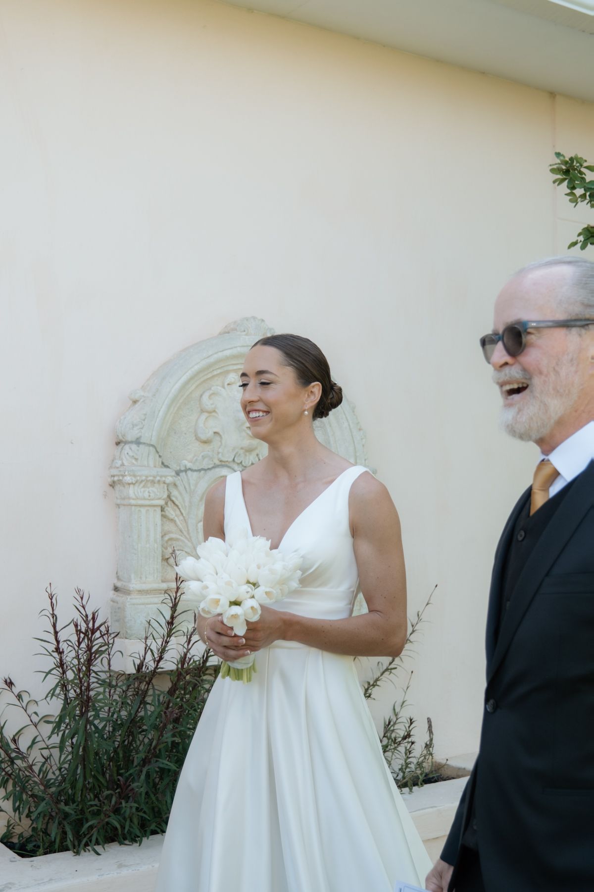 Smiling bride in white V-neck wedding gown holds white floral bouquet beside older gentleman outdoors.