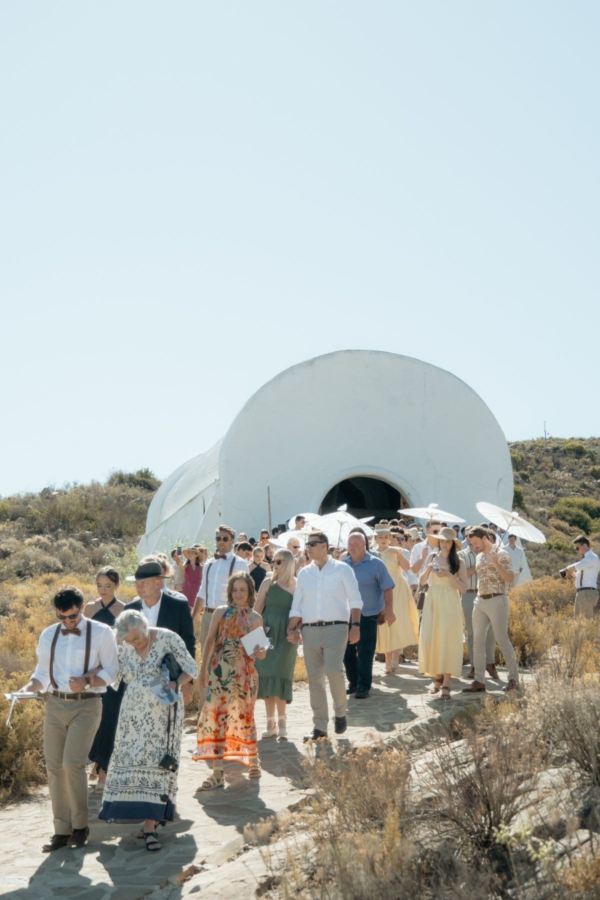 Wedding guests walking away from a white dome chapel in a sunny arid landscape after an outdoor ceremony.