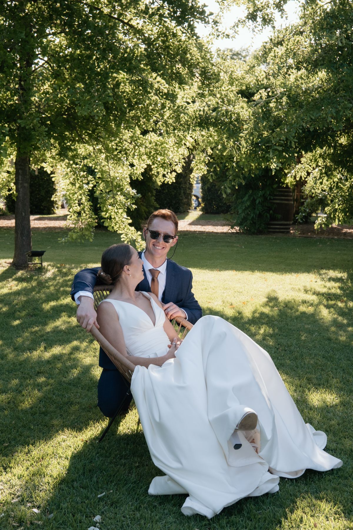 Smiling groom in navy suit sits with bride in white gown on lush green grass beneath shady trees.