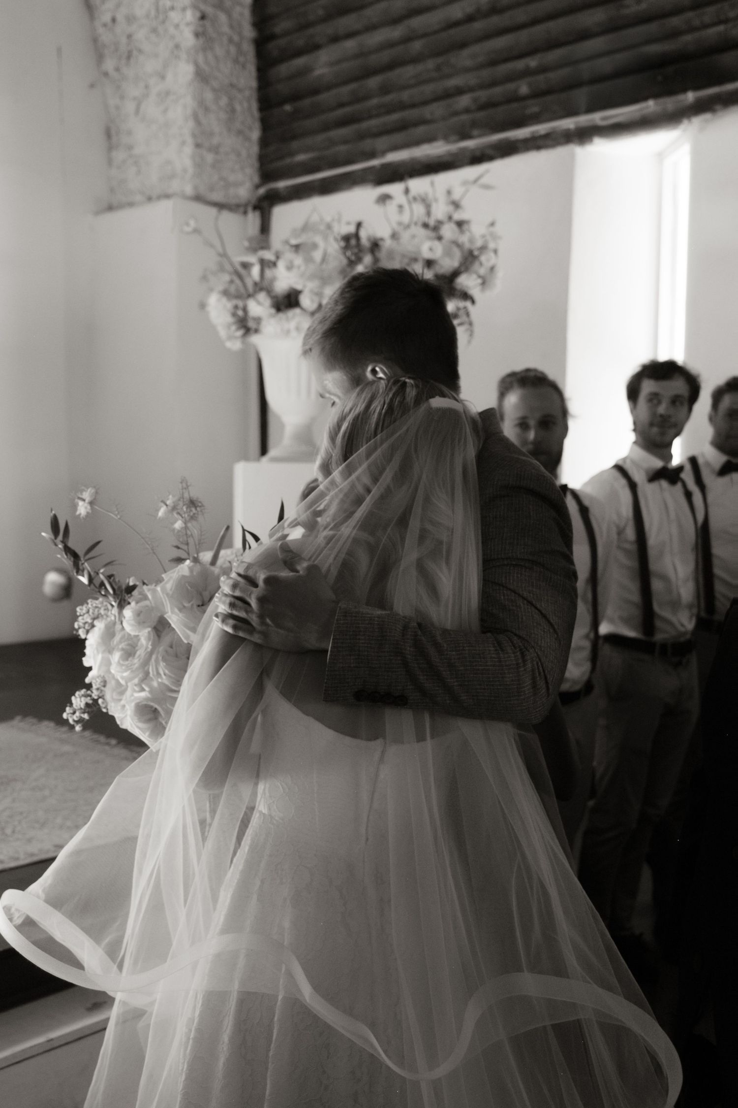 Bride and groom share first dance embrace in black and white wedding photo with guests watching.