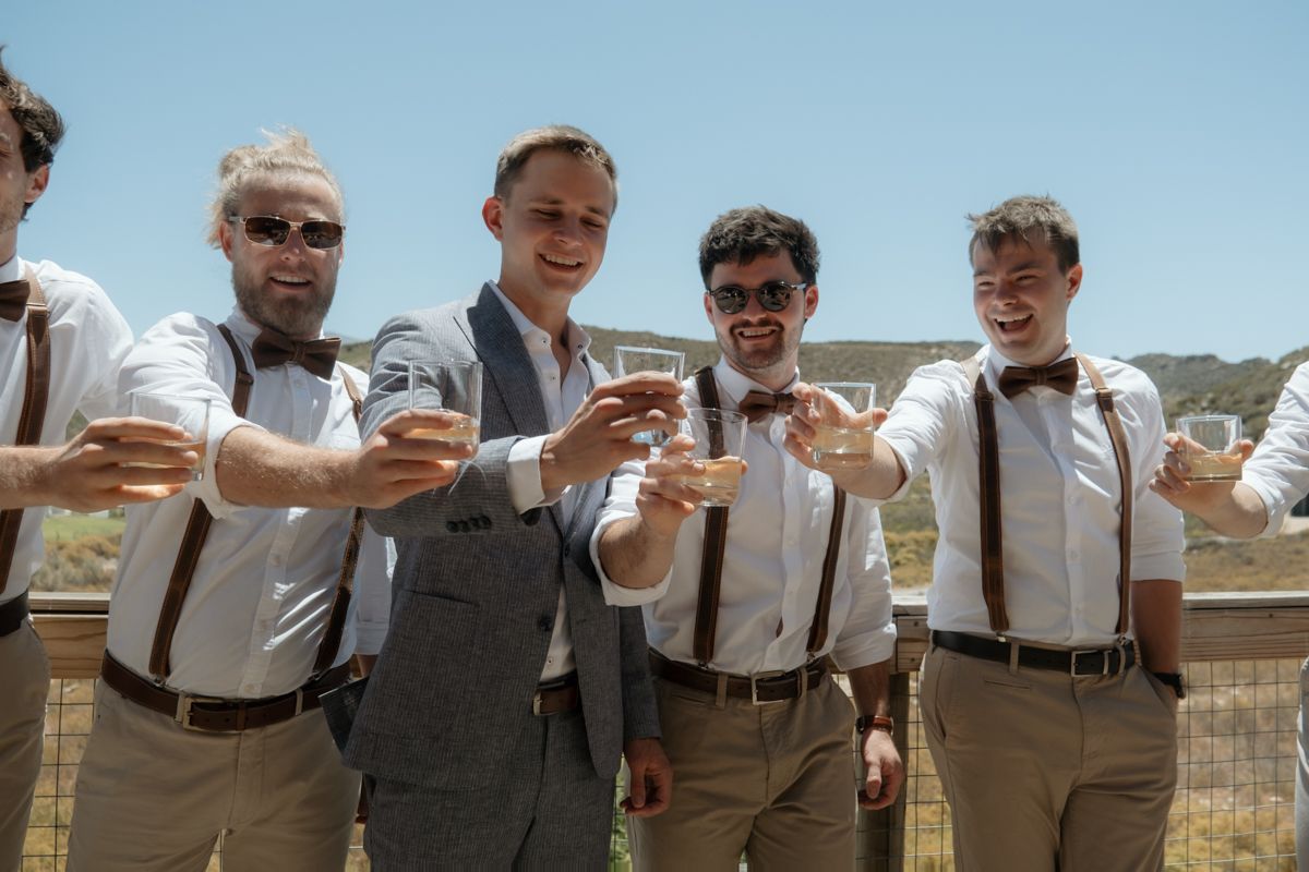 Five groomsmen in white shirts, suspenders, and bow ties toasting with drinks outdoors on a sunny day.