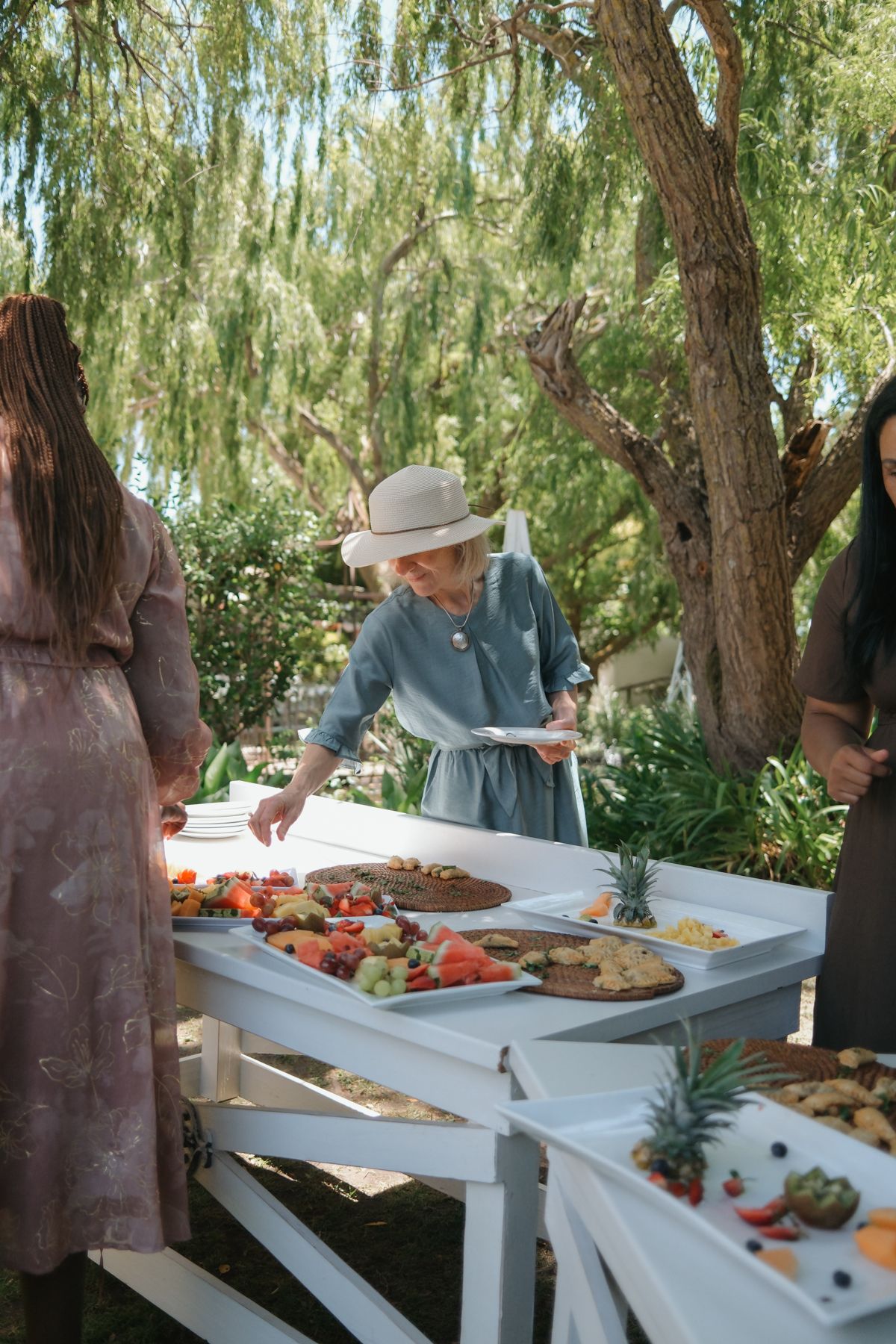 Elderly woman in blue dress and white hat selecting food at an outdoor wedding reception buffet table with fresh fruit.