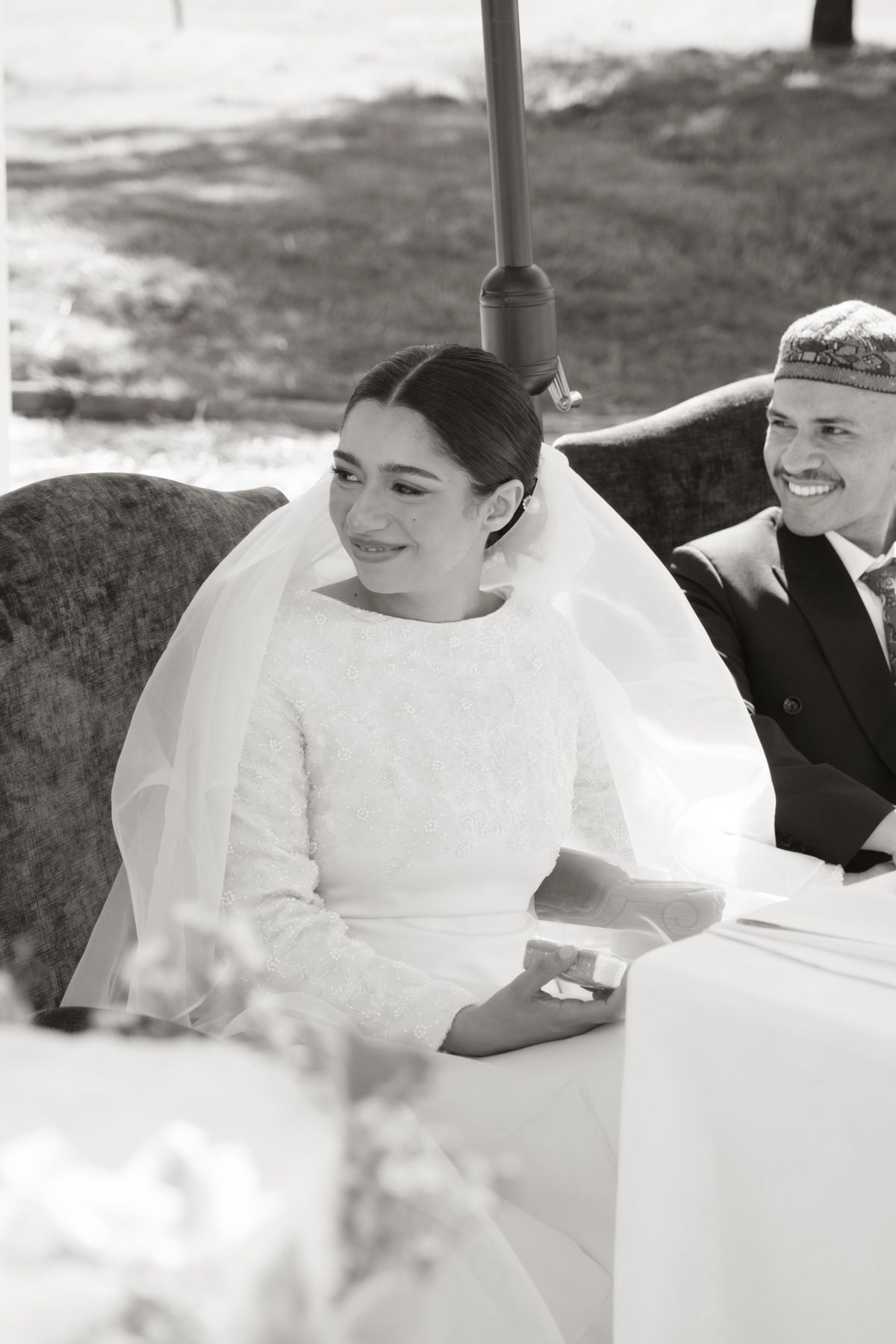 Black and white photo of a smiling bride in veil seated beside a laughing groom at their wedding.