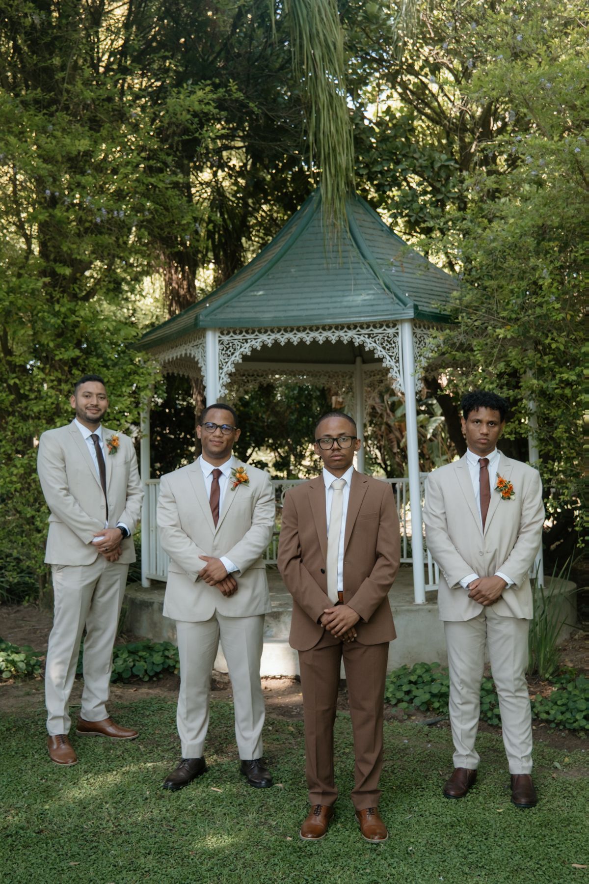 Four groomsmen in beige and brown suits with floral boutonnieres pose in front of a white garden gazebo.