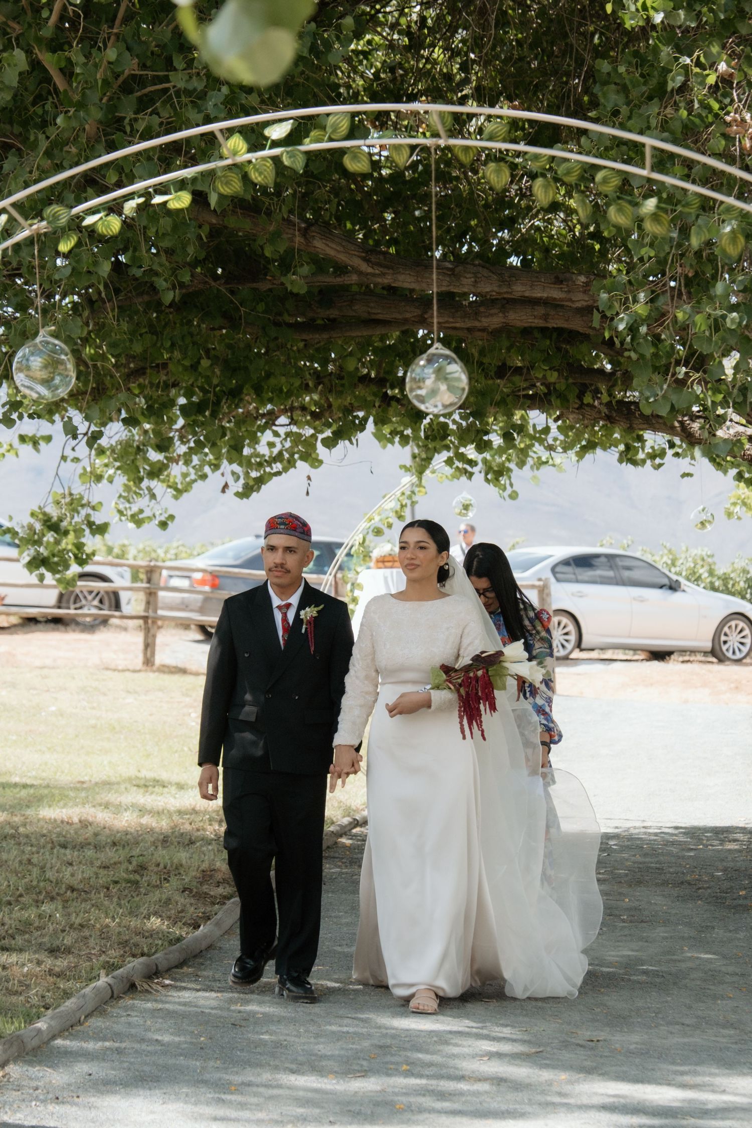 Bride in white gown walks with groom in black suit under floral arch at outdoor wedding ceremony.