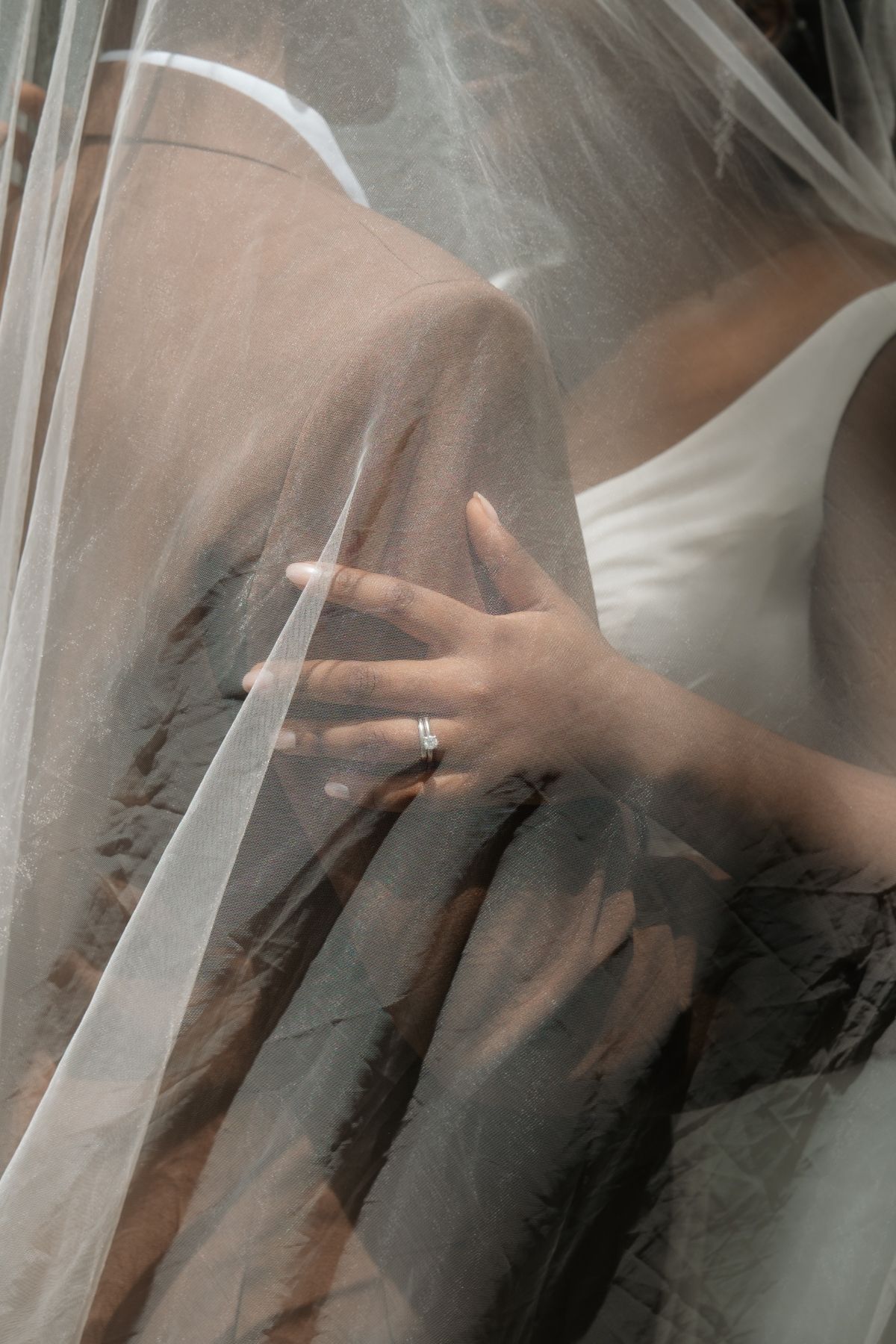 Close-up of bride's hand with diamond engagement ring resting on groom's lap beneath a sheer wedding veil.