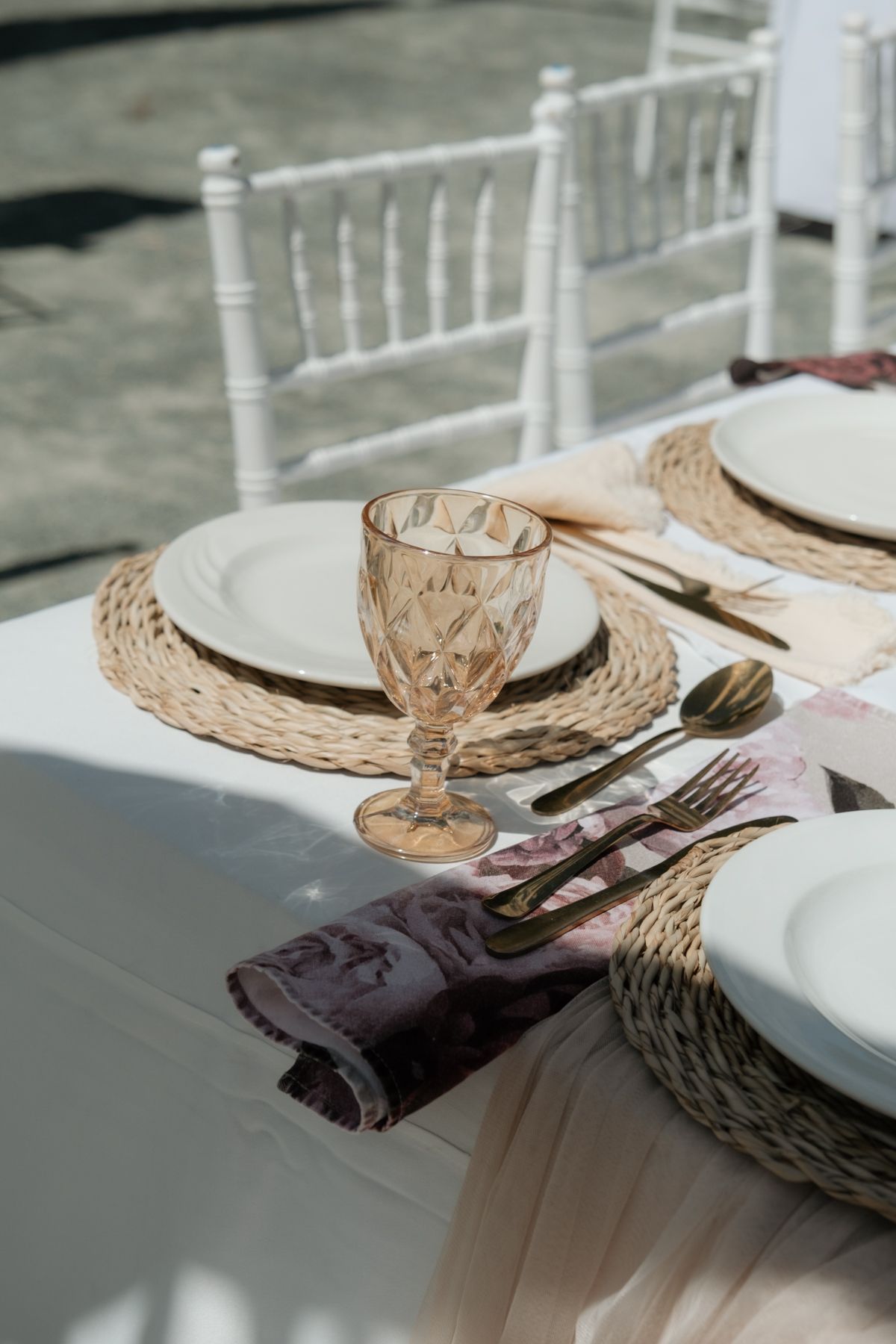 Elegant outdoor wedding table setting with woven placemats, amber glassware, gold cutlery, and white chiavari chairs.