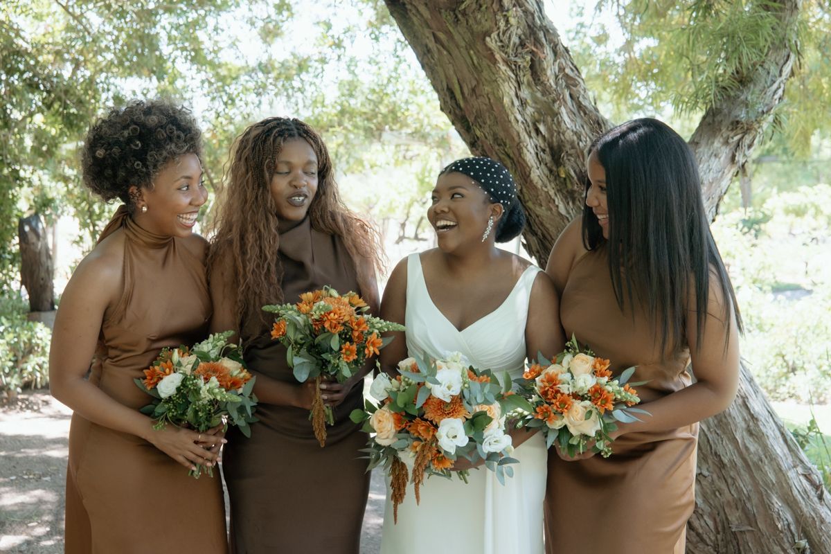 Bride and three bridesmaids in brown dresses laugh joyfully while holding orange and white floral bouquets under a tree.