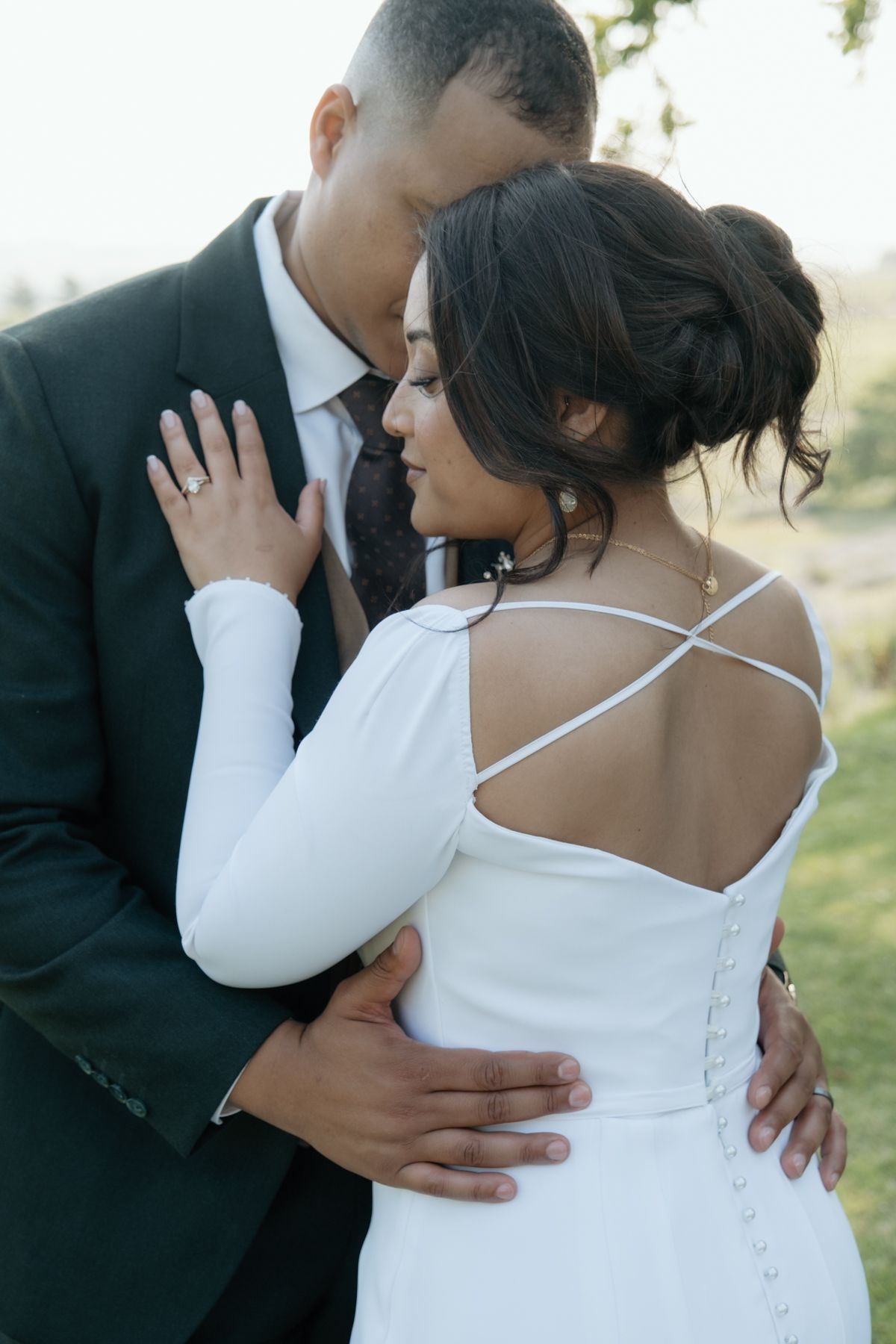 Groom tenderly embraces bride from behind, showcasing her elegant backless white wedding gown.