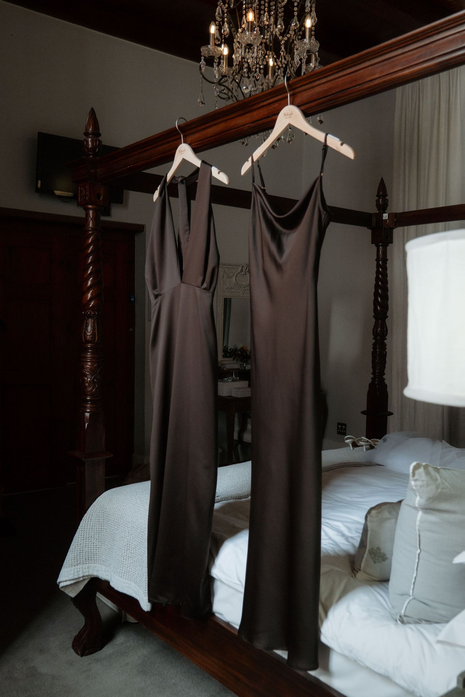 Two dark olive satin bridesmaid dresses hanging on wooden hangers in an elegant bedroom with chandelier.