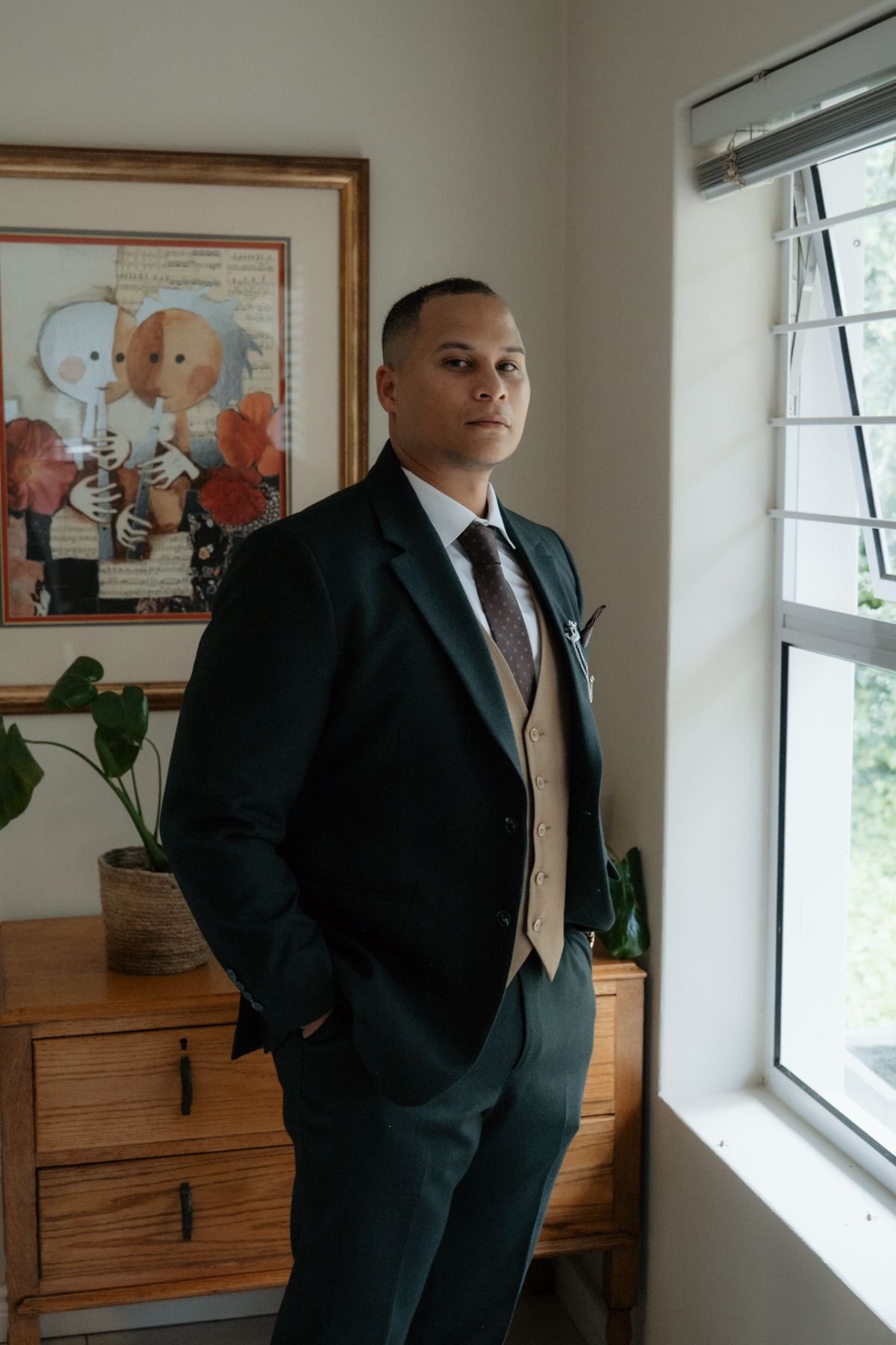 Dapper groom in dark green suit with tan vest poses thoughtfully beside a window before wedding.