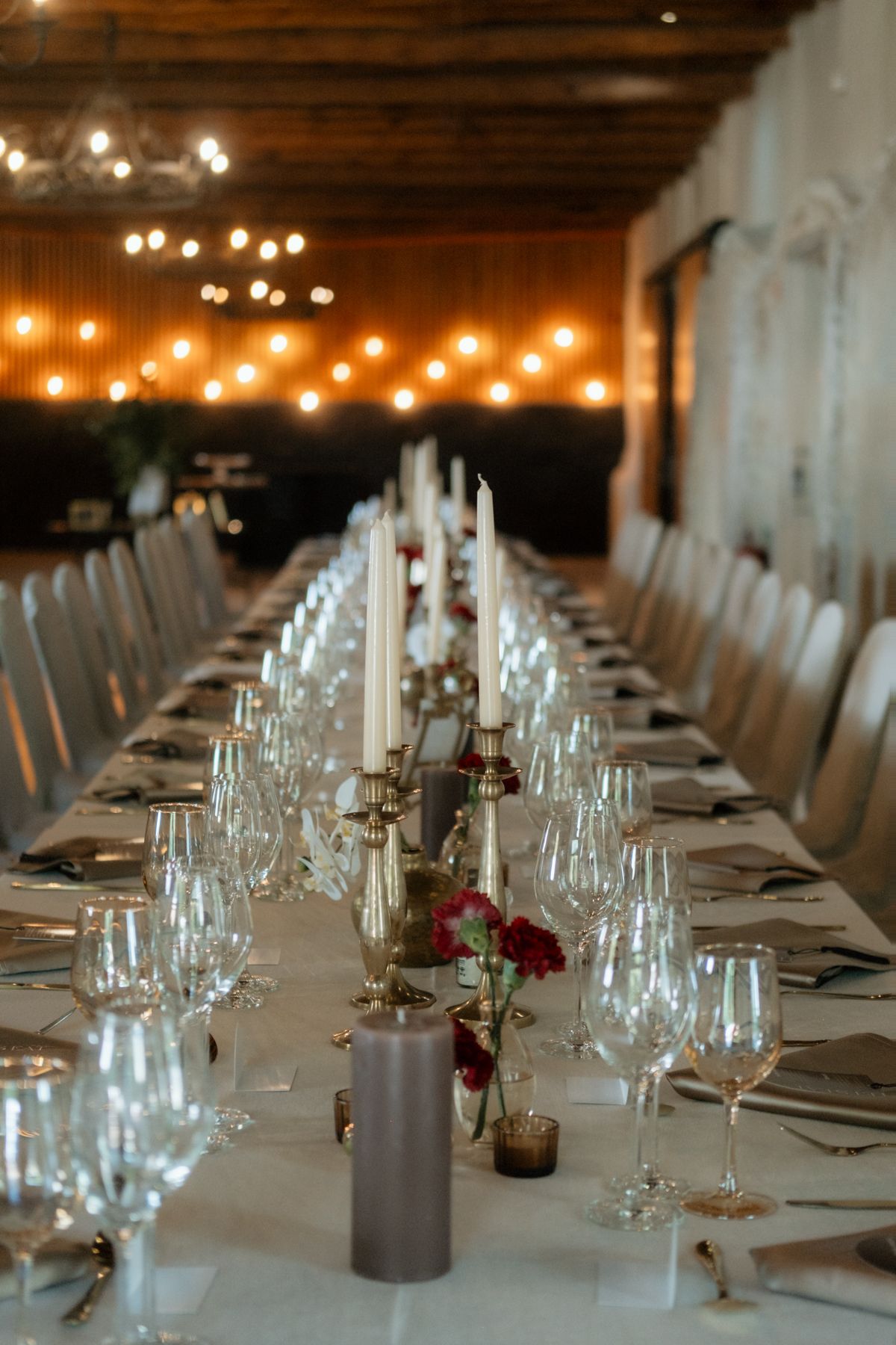 Elegant wedding reception table set with white candles, crystal glasses, red floral centerpieces, and warm string lights.