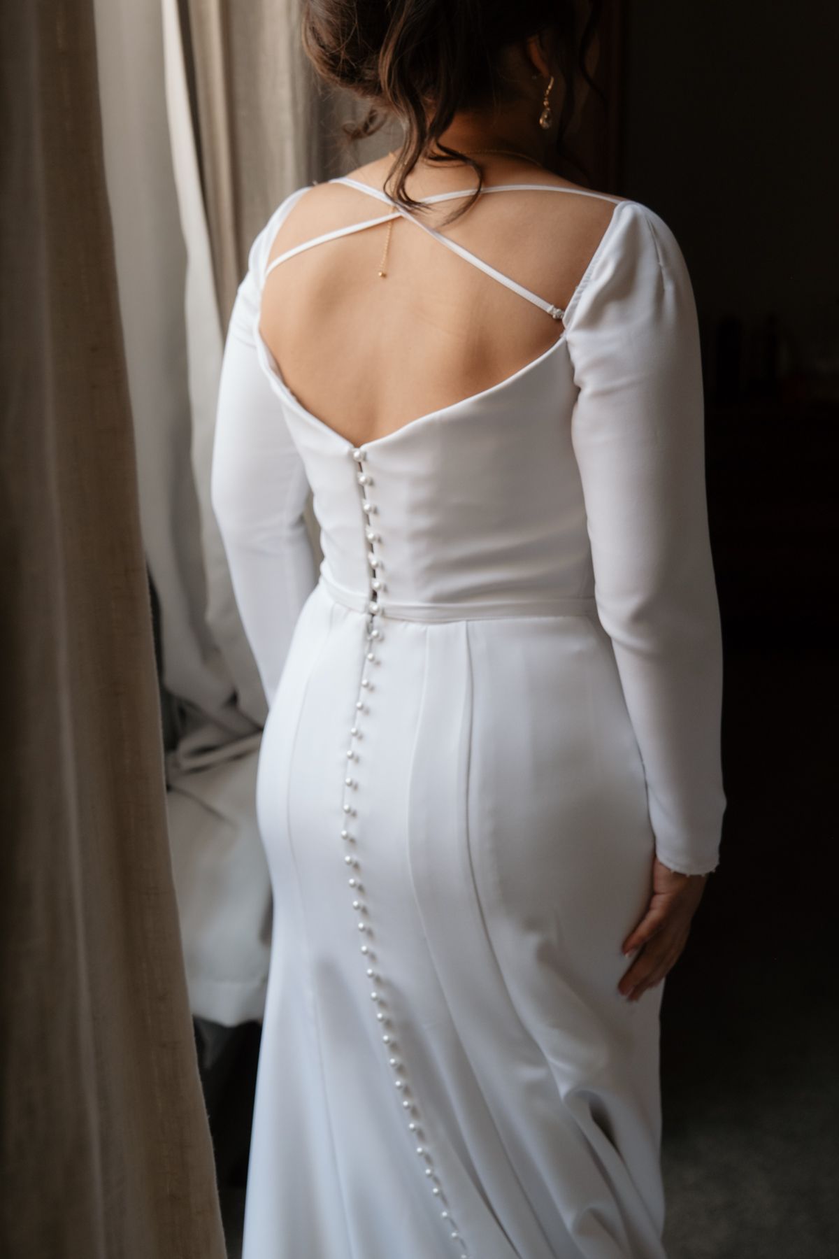 Bride in elegant white long-sleeve wedding gown with button back detail and criss-cross straps, standing by window.