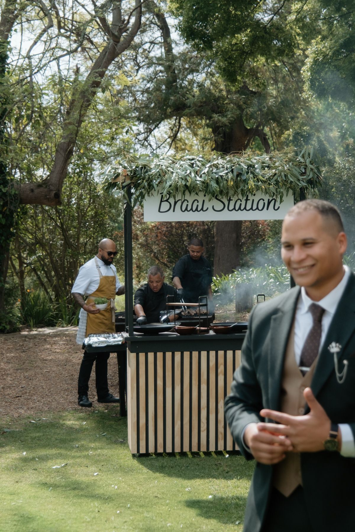 Outdoor braai station with chefs cooking at wedding reception surrounded by lush trees with smiling guest in foreground.