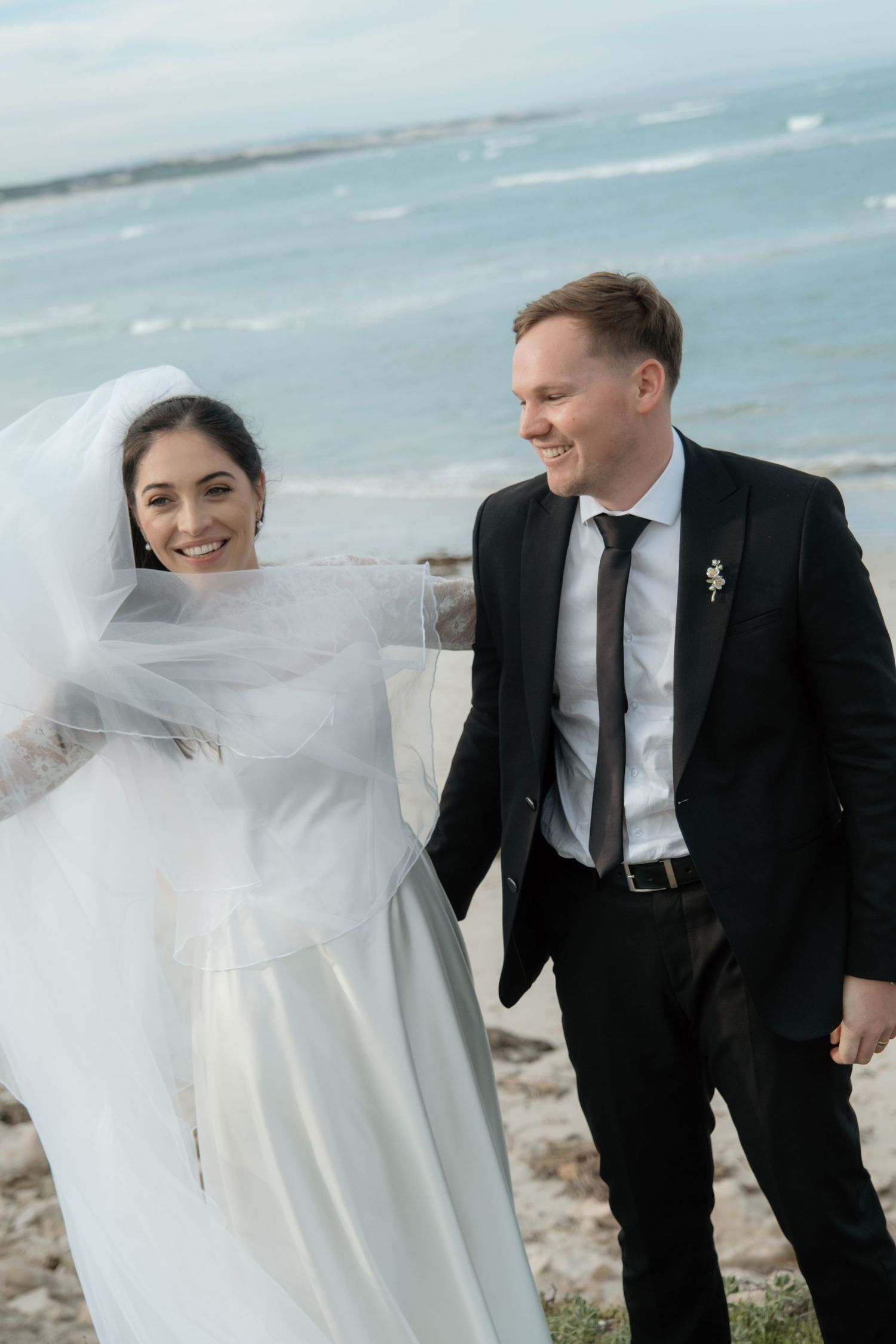 Smiling bride in white gown and veil walks with groom in black suit near ocean on their wedding day.