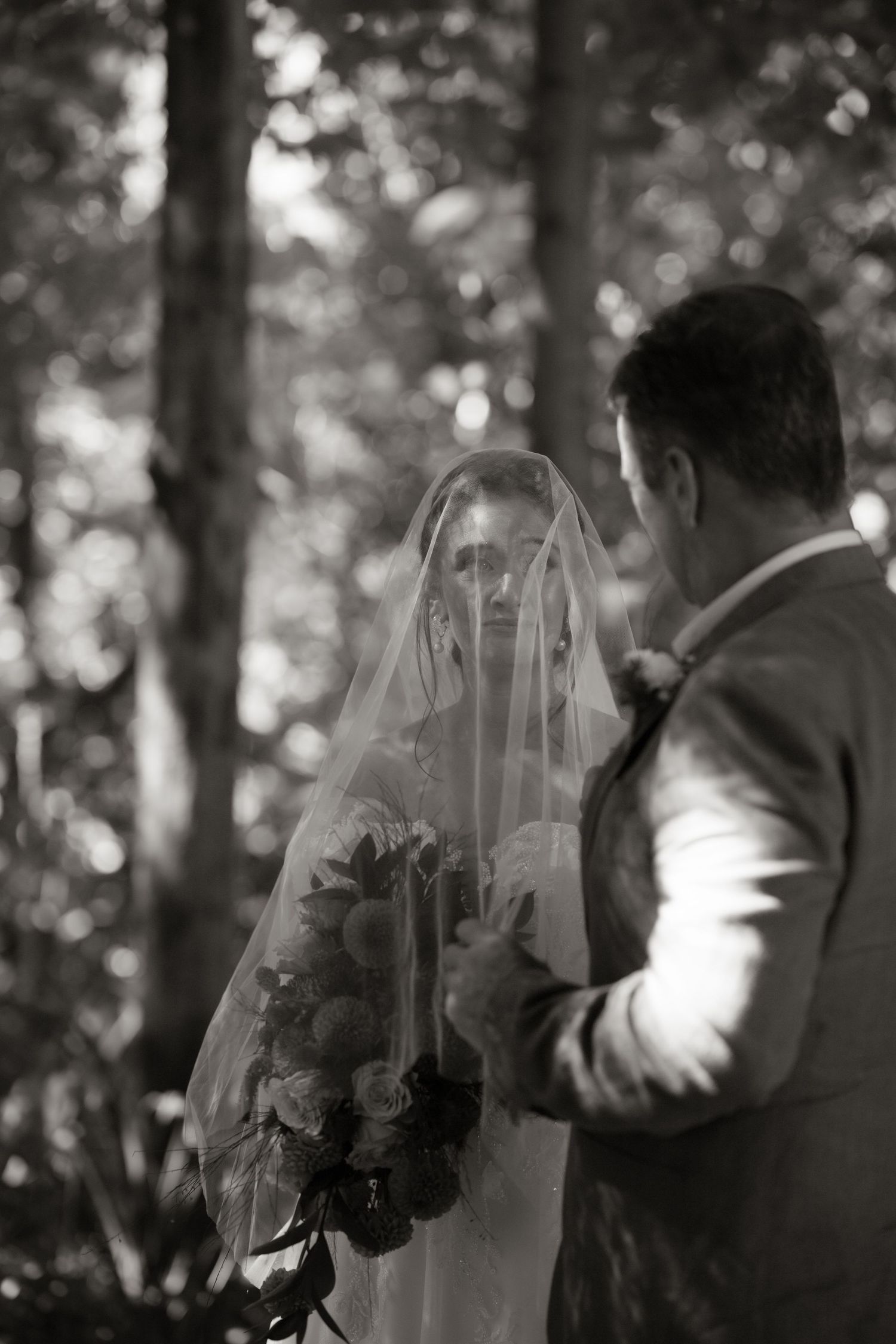 Black and white photo of groom lifting bride's veil during an intimate first look in the forest.