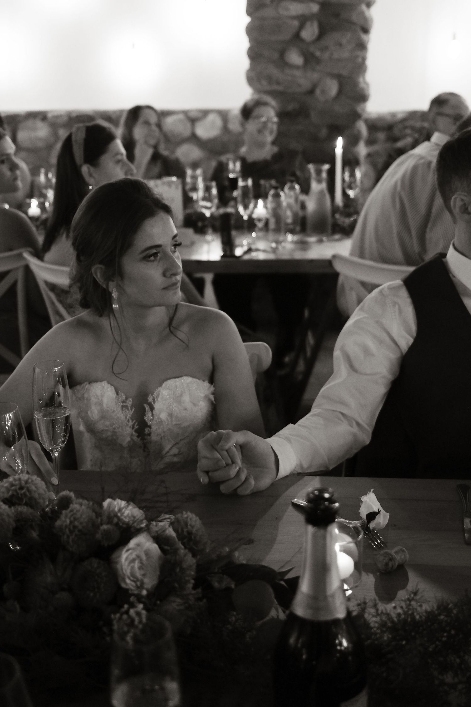 Black and white photo of bride in strapless lace gown holding grooms hand at reception table.