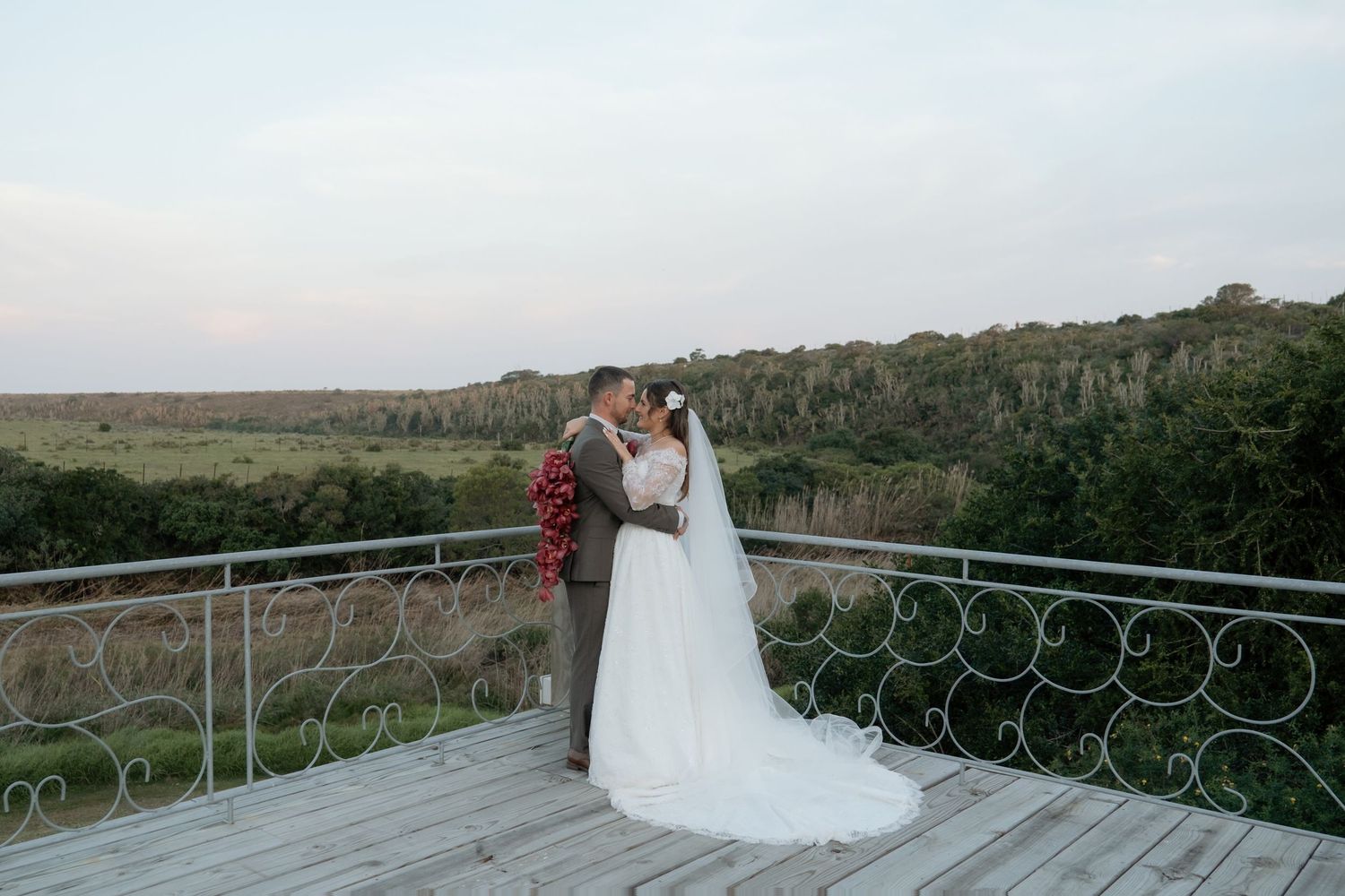 Bride and groom share a romantic kiss on a scenic outdoor terrace overlooking lush green landscape at sunset.