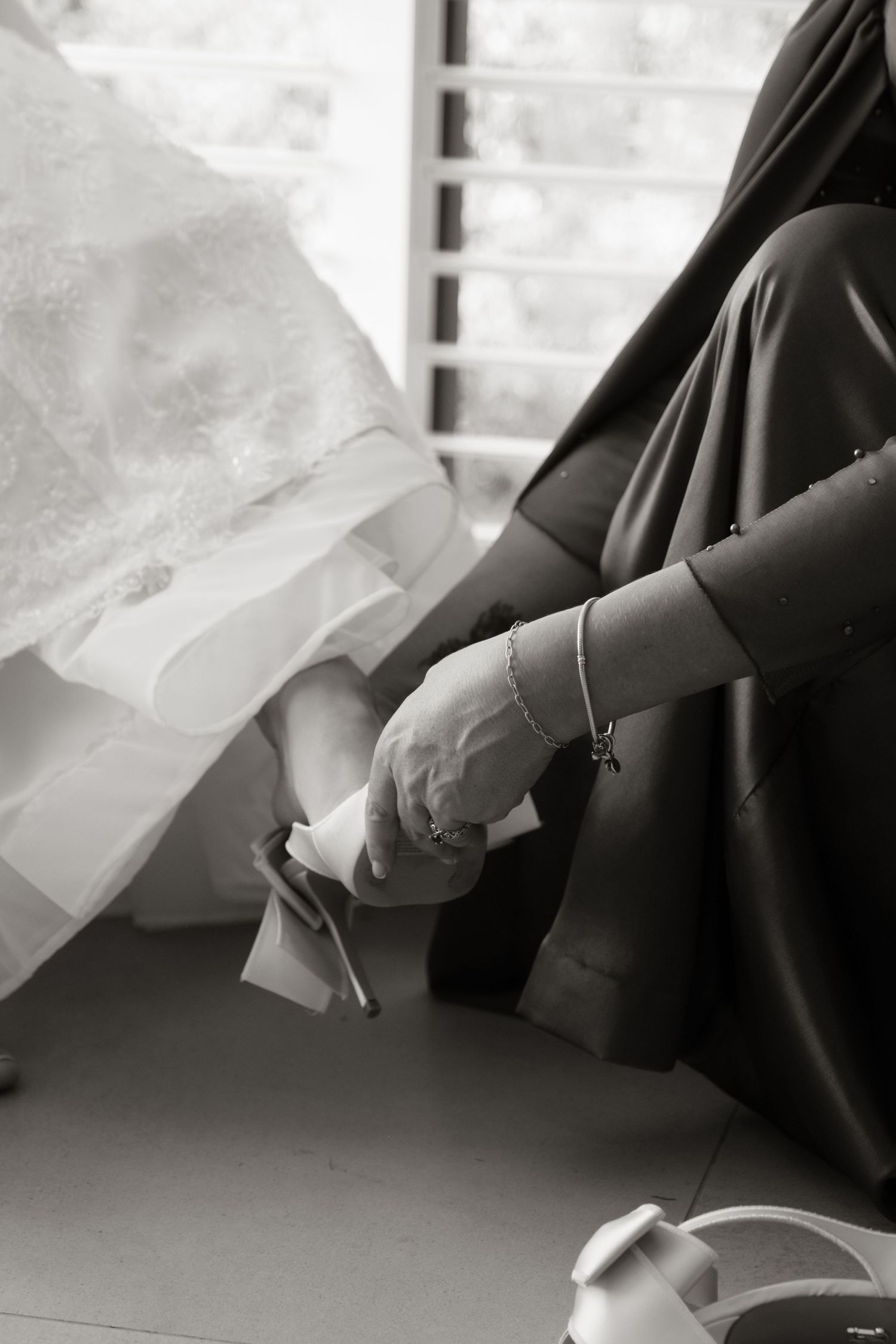 Black and white photo of bride getting shoes put on by helper in dark dress.