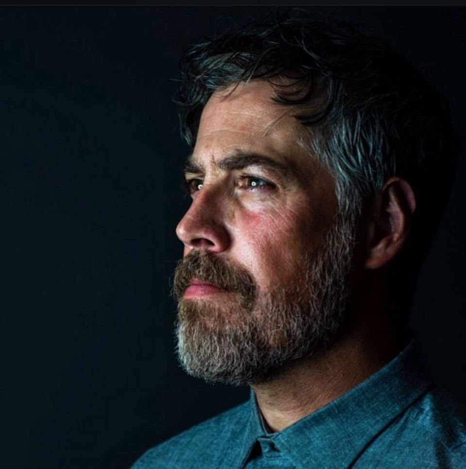 Dramatic side profile portrait of a bearded man in blue shirt against dark background with moody lighting.