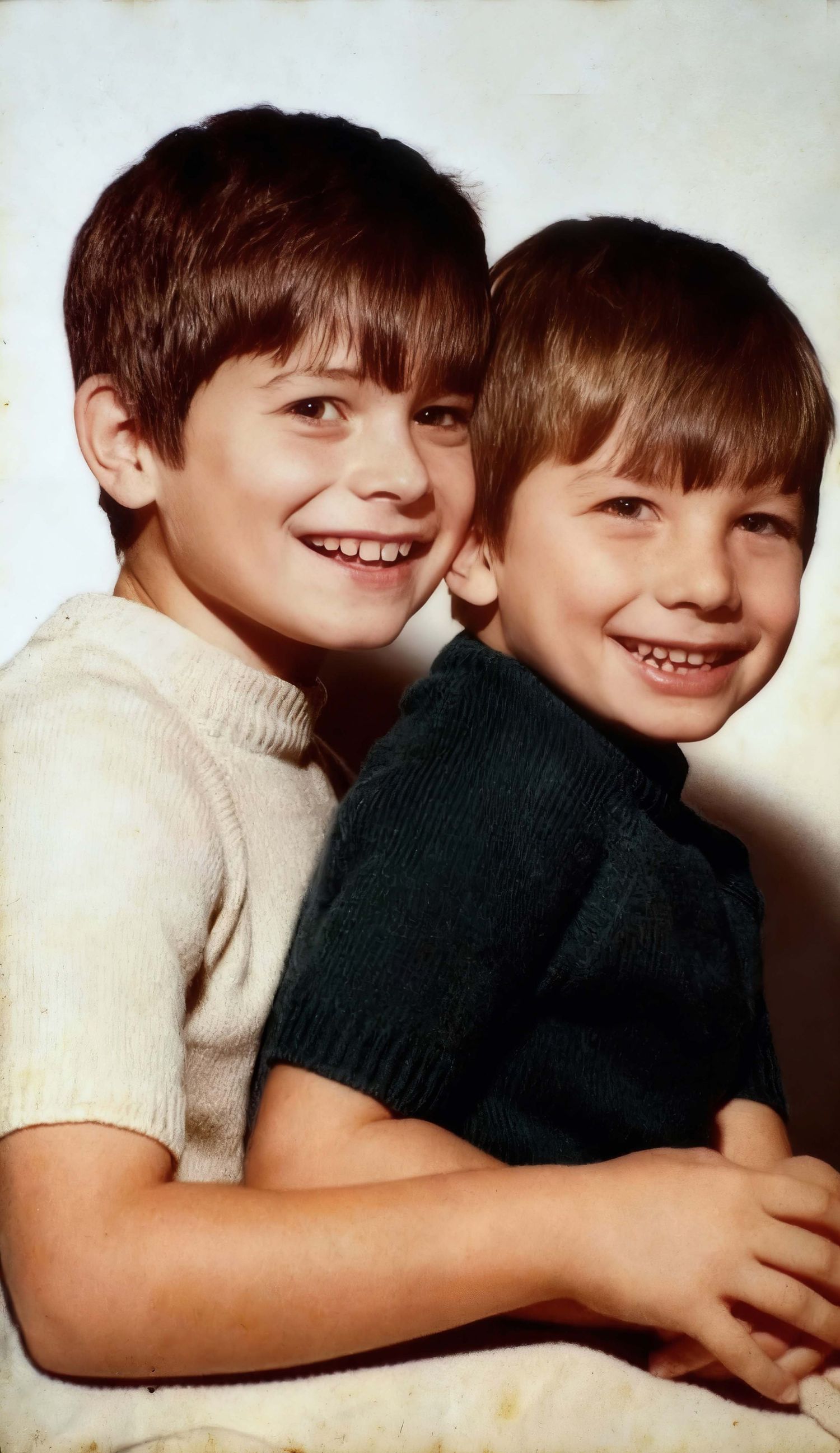 Two children in white and black sweaters share a warm embrace while smiling at the camera.